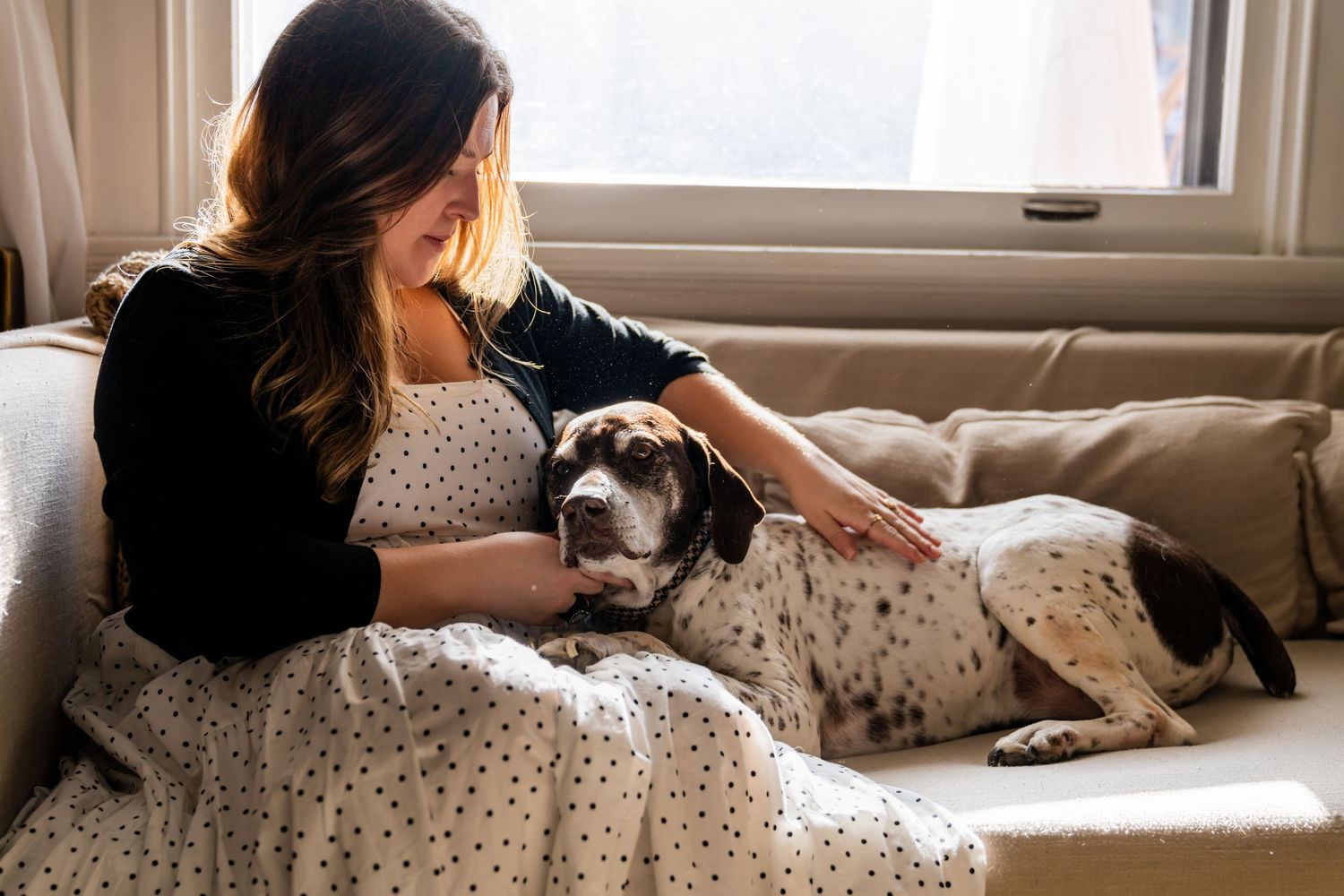 Person in white polka dot dress sits on couch petting their spotted black and white dog in sunlit room during an end of life dog session