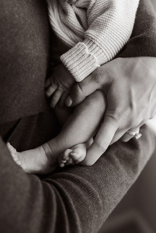 Black and white close-up photo of hands gently holding a sleeping baby's tiny feet in a knitted sweater.