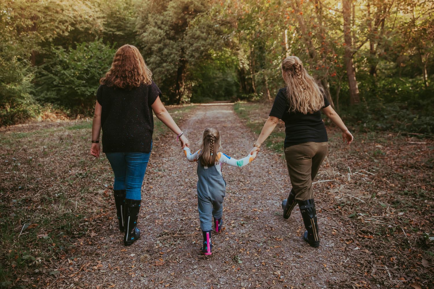 Three people walking hand in hand down a gravel path through nature during golden hour. During a family photoshoot in High elms
