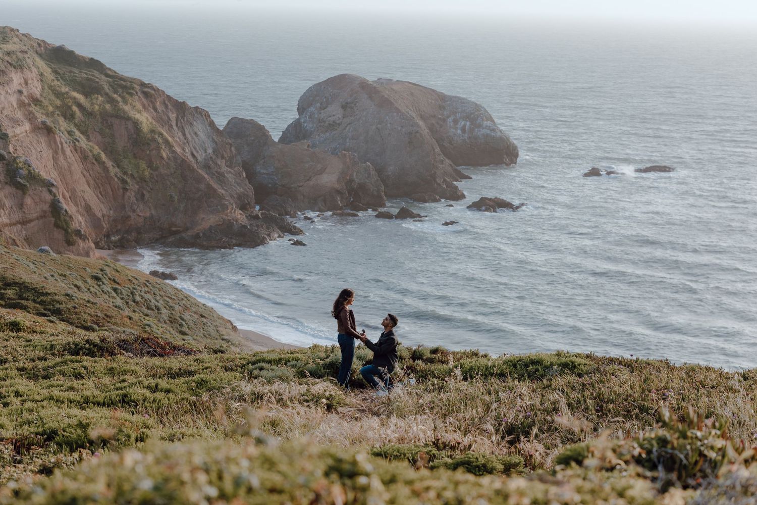 Two people in white dresses dance together on grassy coastal cliffs overlooking dramatic rock formations and ocean views.