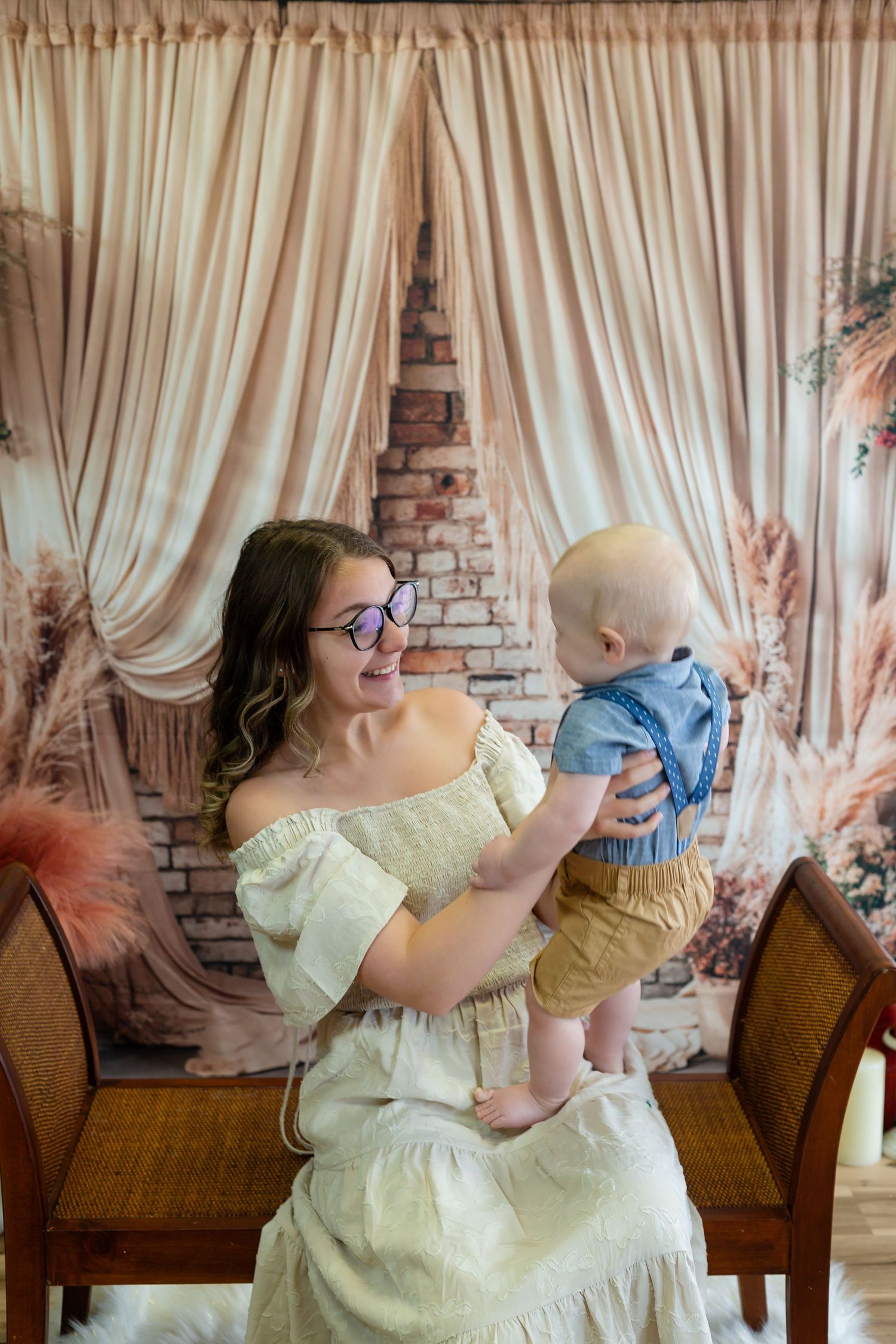In an elegant vintage setting with cream curtains and brick walls, a person in a yellow dress lifts a baby wearing blue.