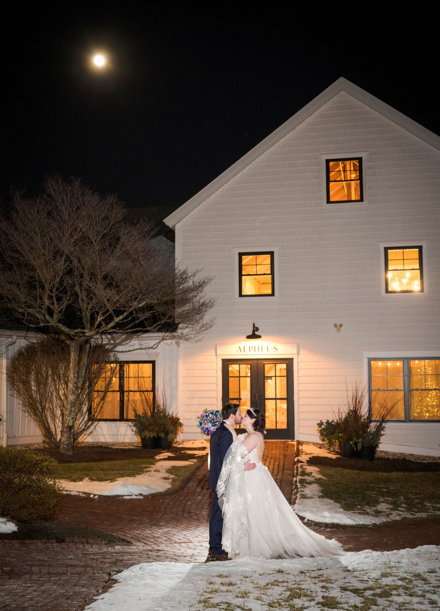 A winter wedding portrait outside a white farmhouse with illuminated windows and a full moon.