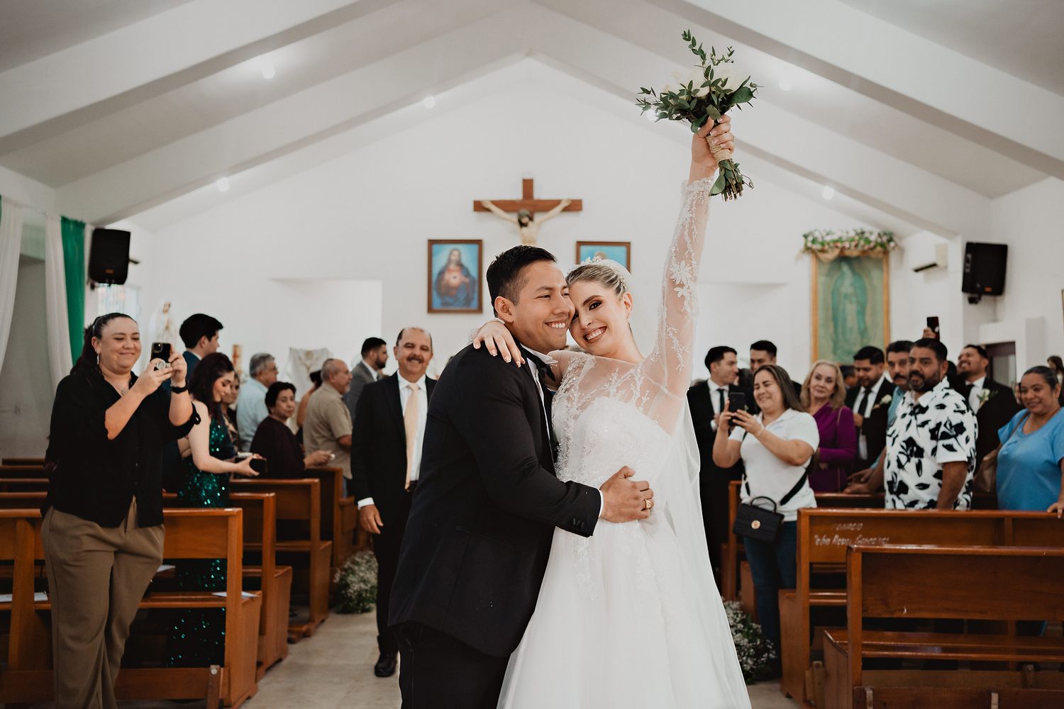 Joyful newlyweds celebrate with raised bouquet during church ceremony as guests look on from wooden pews.