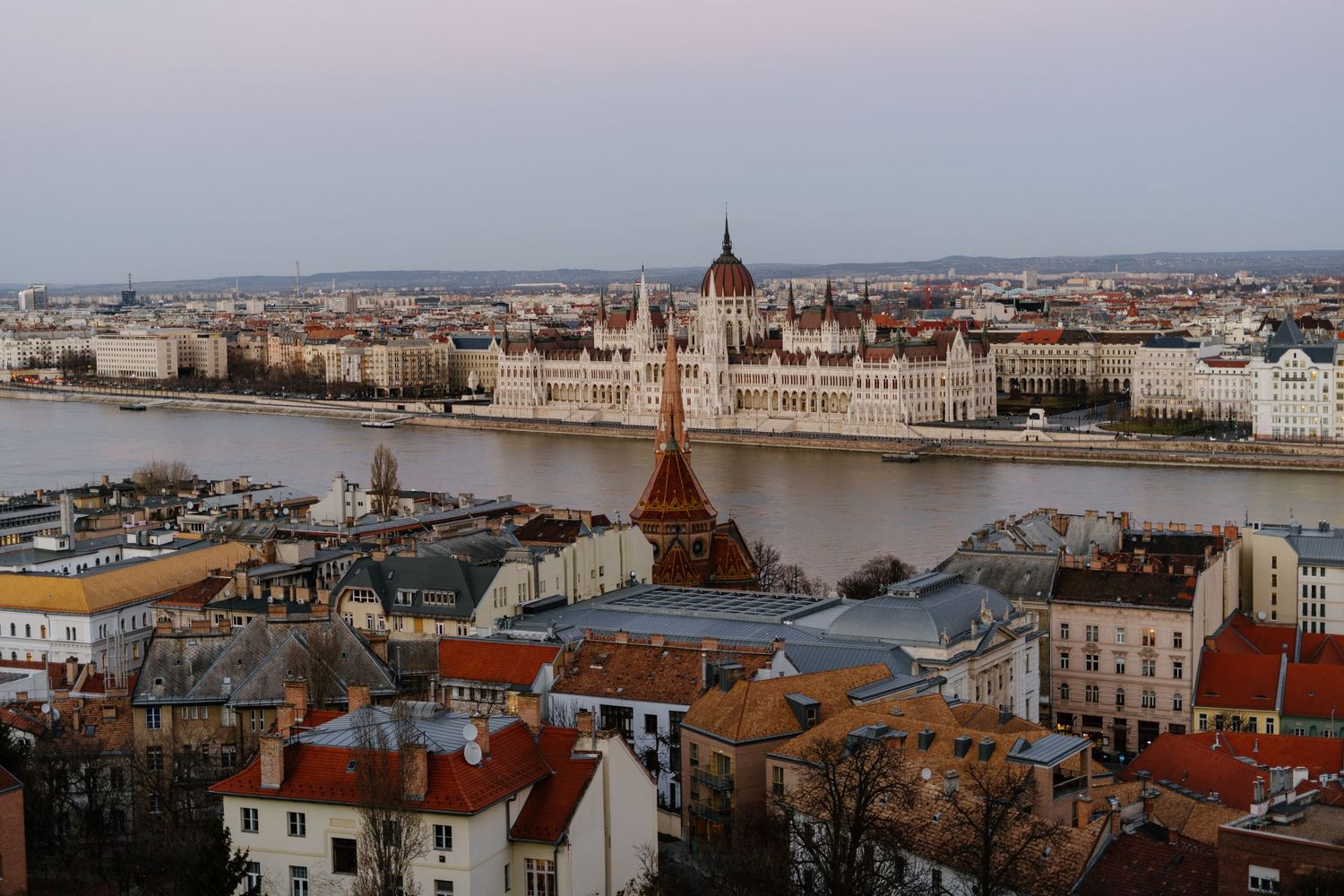 Aerial view of Budapest's Parliament Building along the Danube River with historic rooftops in the foreground at dusk.