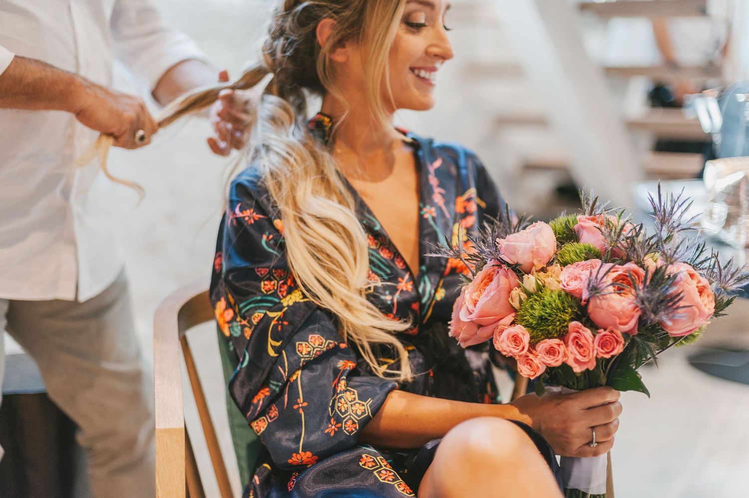 A floral silk robe-clad person smiles while holding a lush pink bouquet during styling preparations.