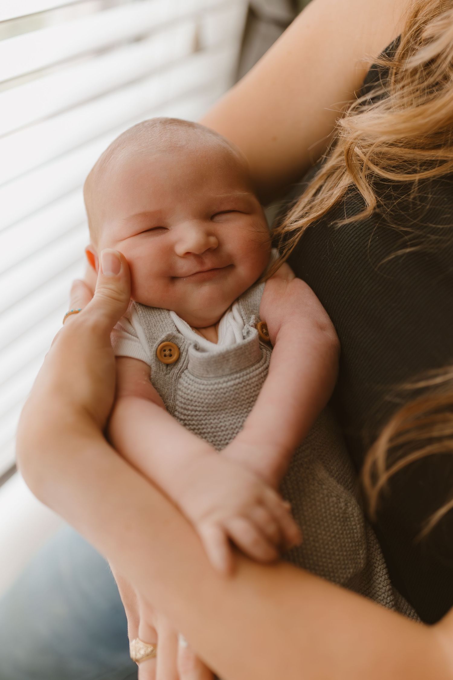 A smiling baby wearing a gray knit romper with wooden buttons is cradled in loving arms by a window.