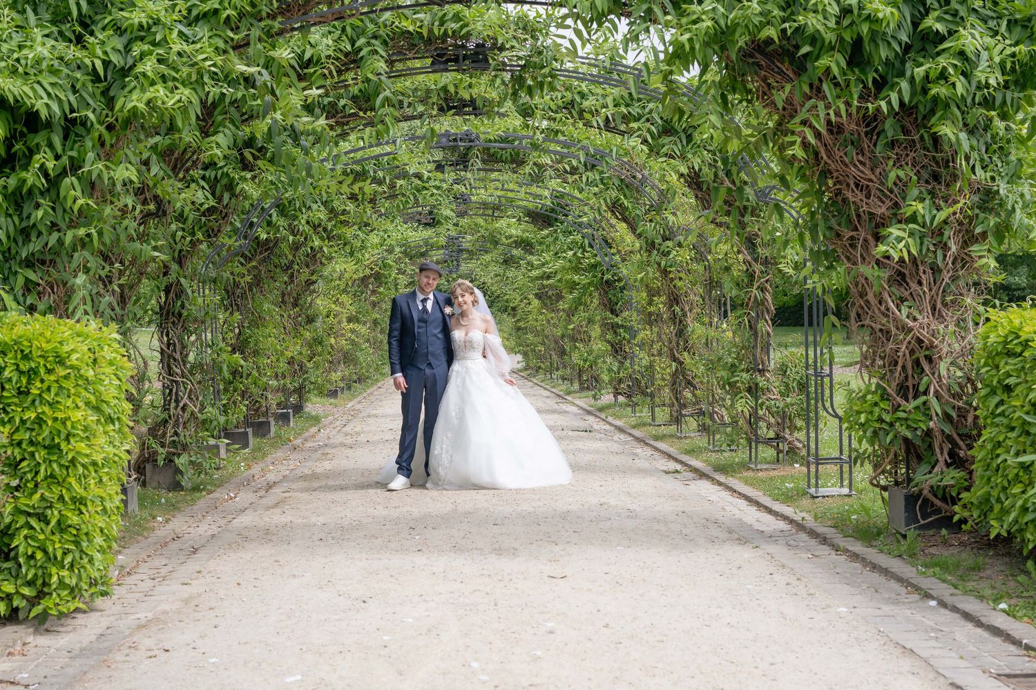 A couple walks down a romantic garden path covered by a lush green archway of plants and vines.