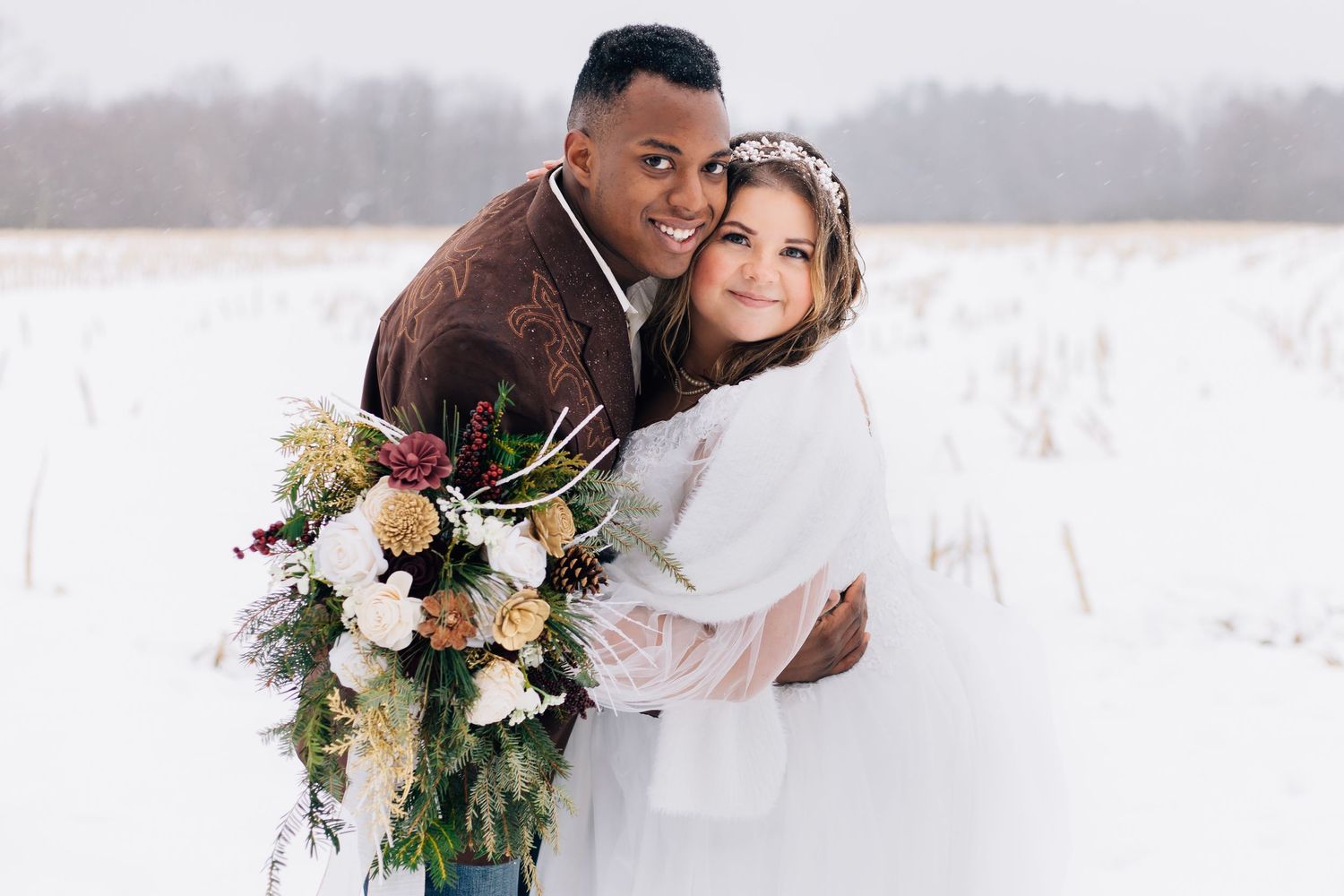 A romantic winter wedding portrait with a couple embracing in a snowy field holding a lush floral bouquet.