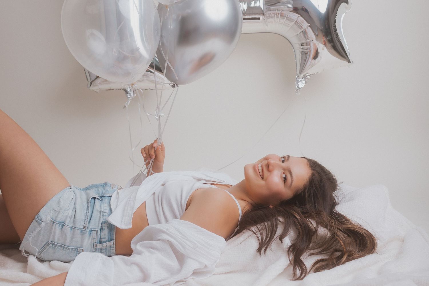 Dreamy portrait with silver balloons and soft white bedding creating ethereal atmosphere.
