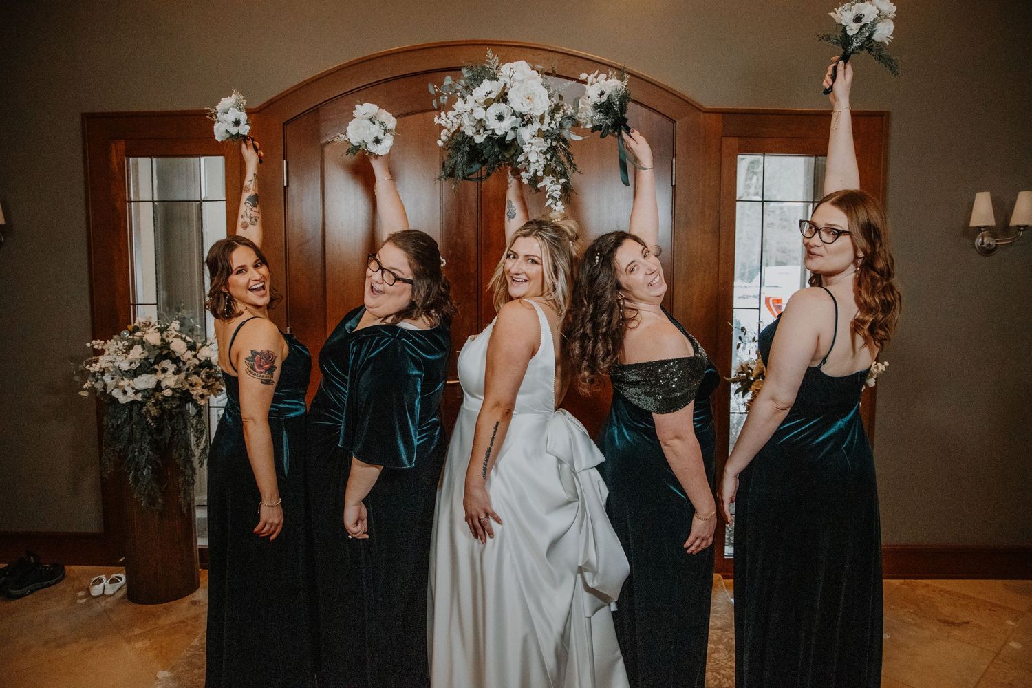 Bride and bridesmaids in elegant green velvet dresses pose together under floral arch at indoor wedding venue.
