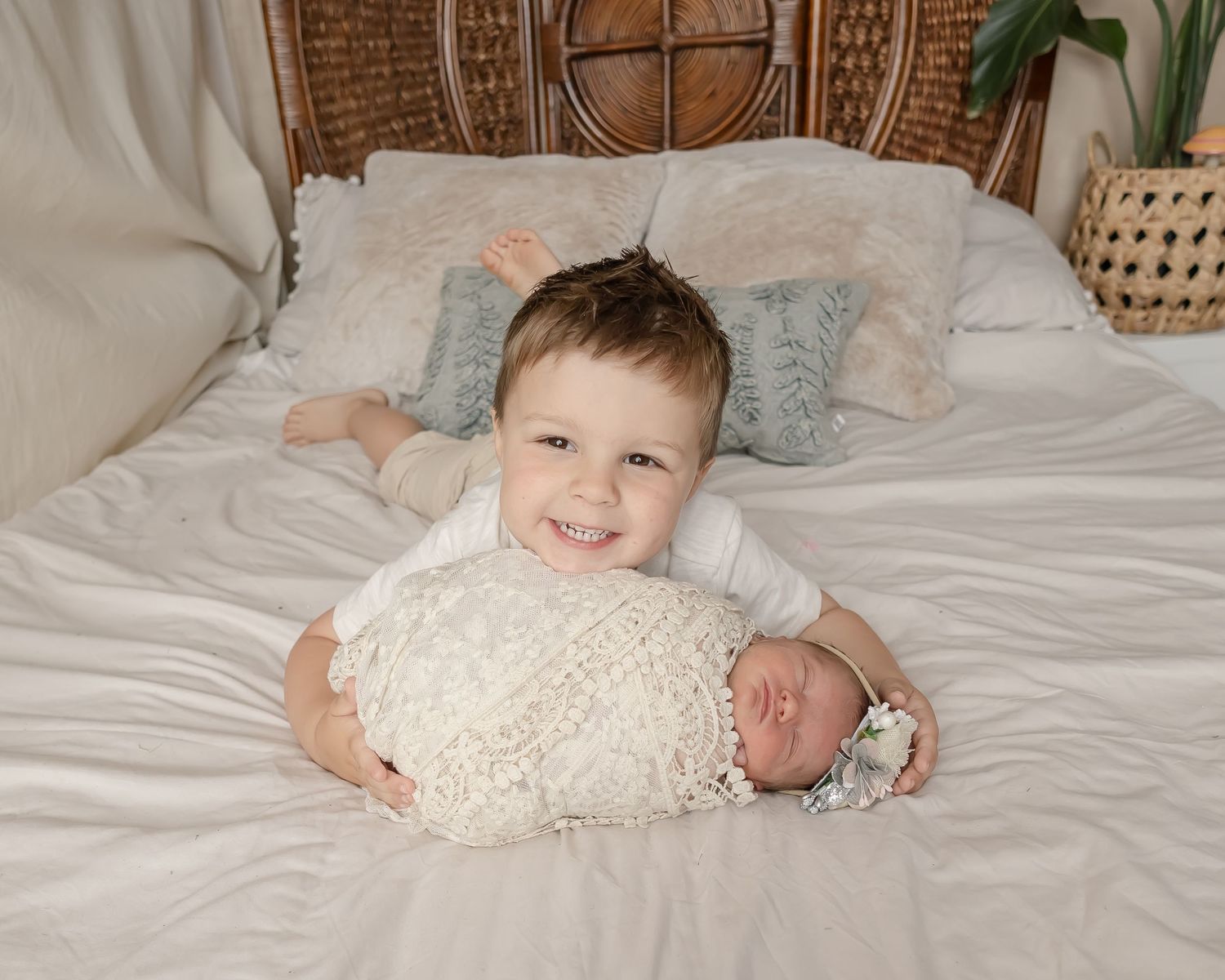 Two siblings on white bedding as older child holds newborn wrapped in lace blanket.