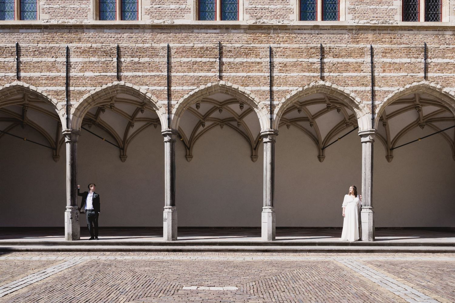 Two figures stand in contrast at opposite ends of an arched stone colonnade in a Renaissance-style courtyard.