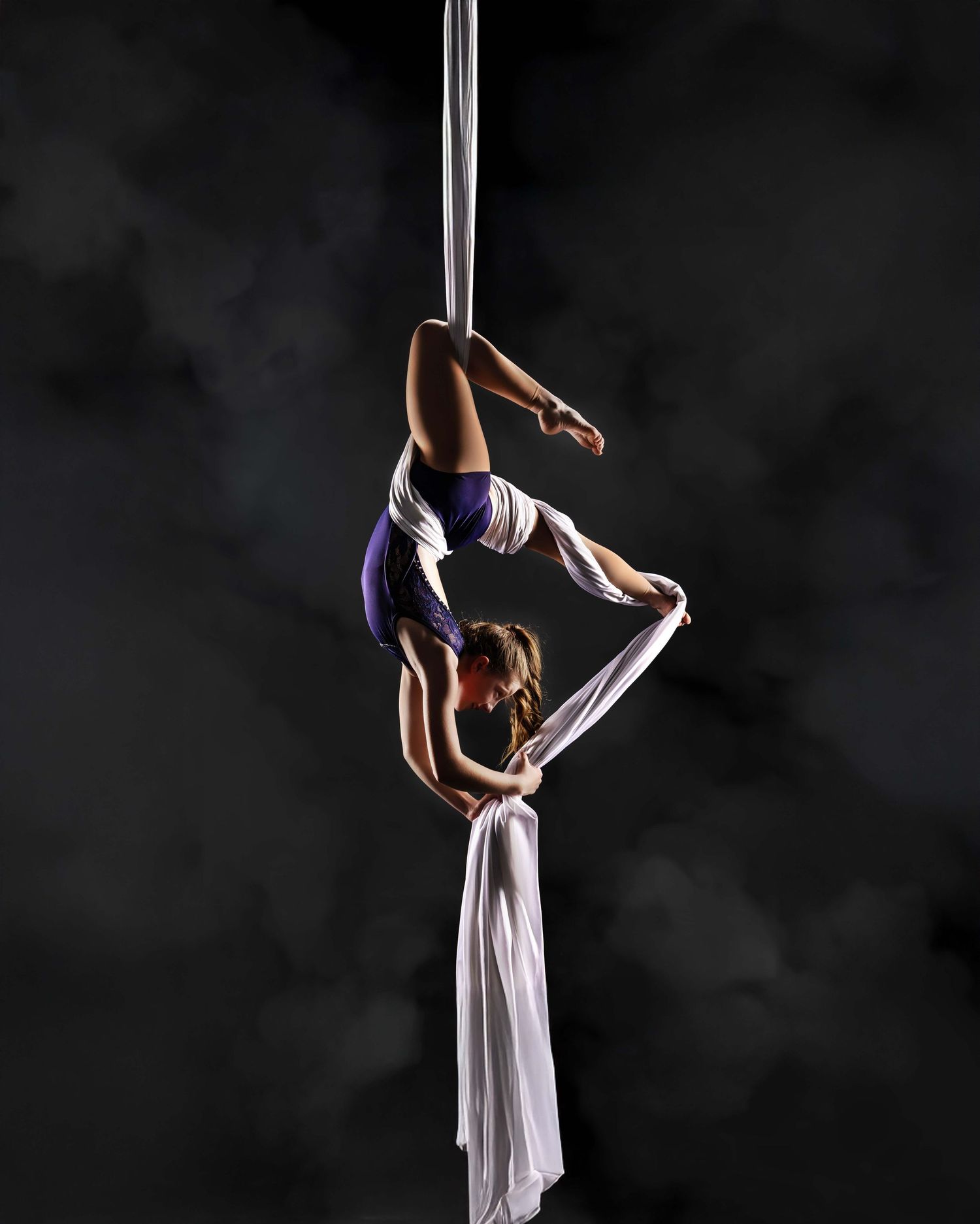 An aerial performer in a red costume gracefully suspends from white silk ribbons against a dark background.