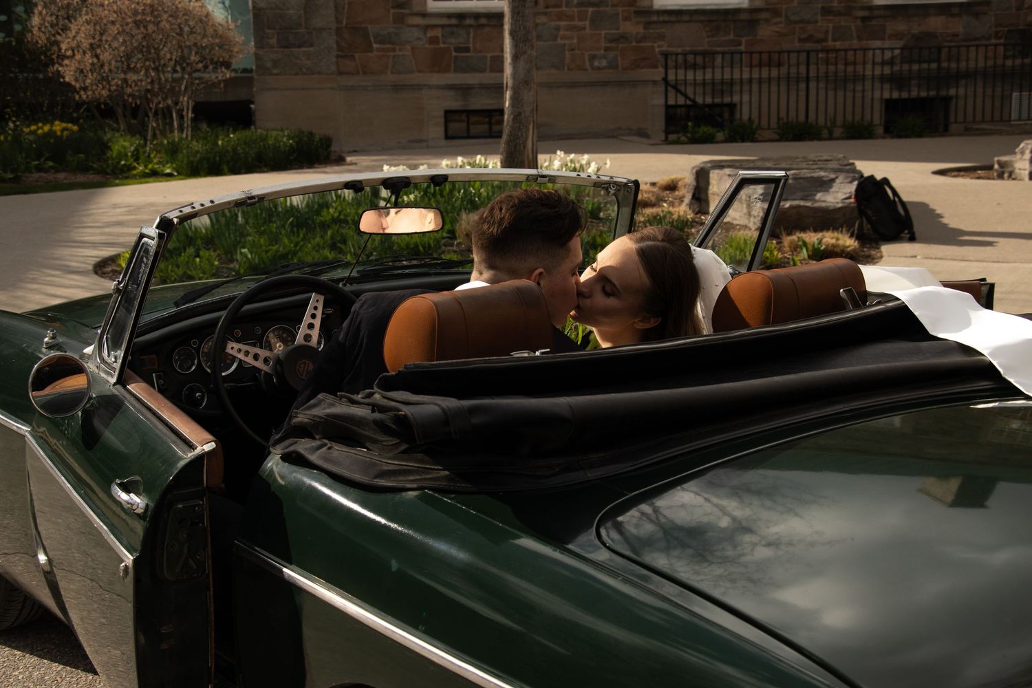A couple is seen from behind, sitting in a dark green convertible car, sharing a kiss. The woman is wearing a wedding dr