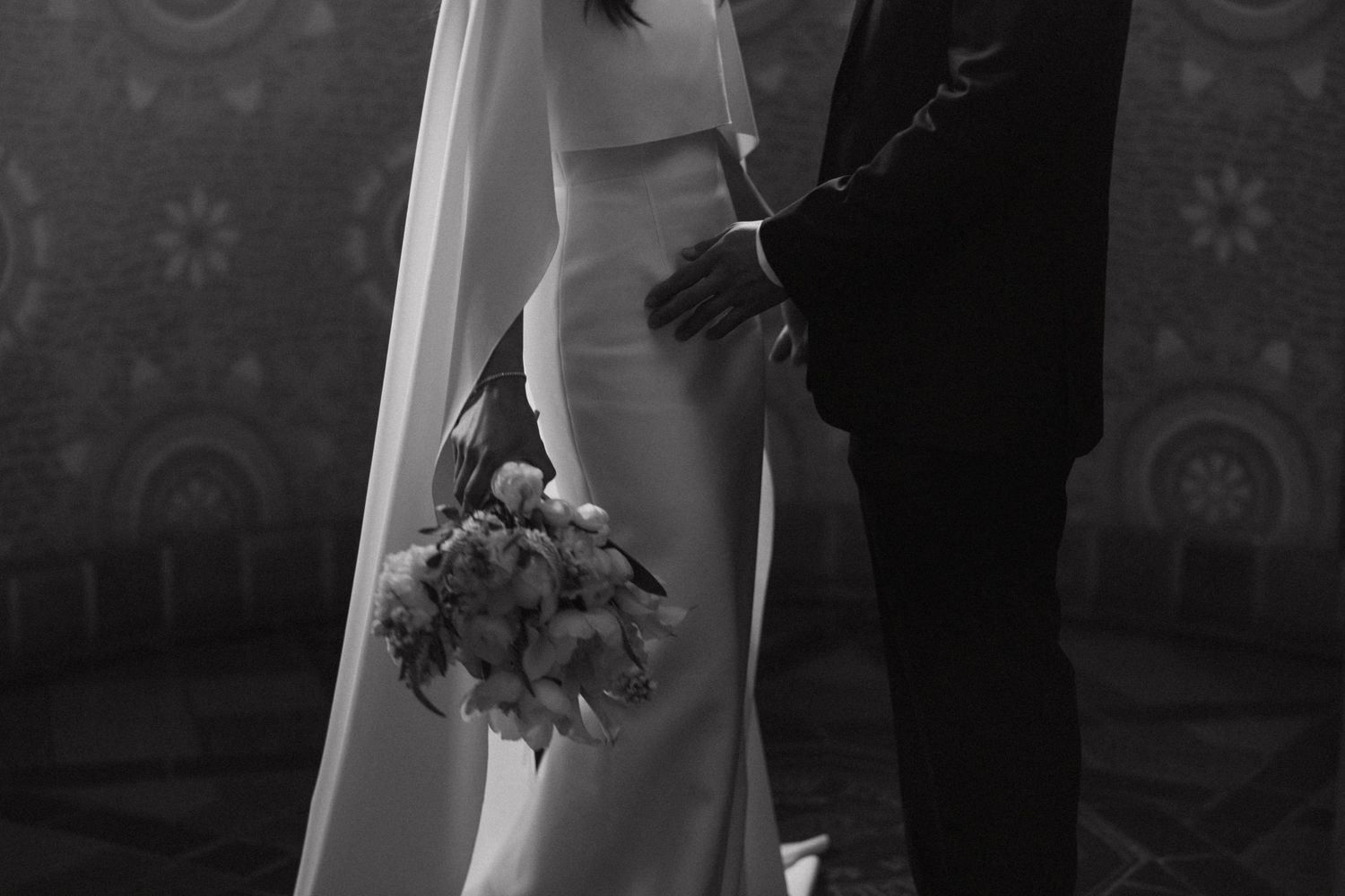 Black and white close-up of bride holding a bouquet while groom holds her hand at the altar.