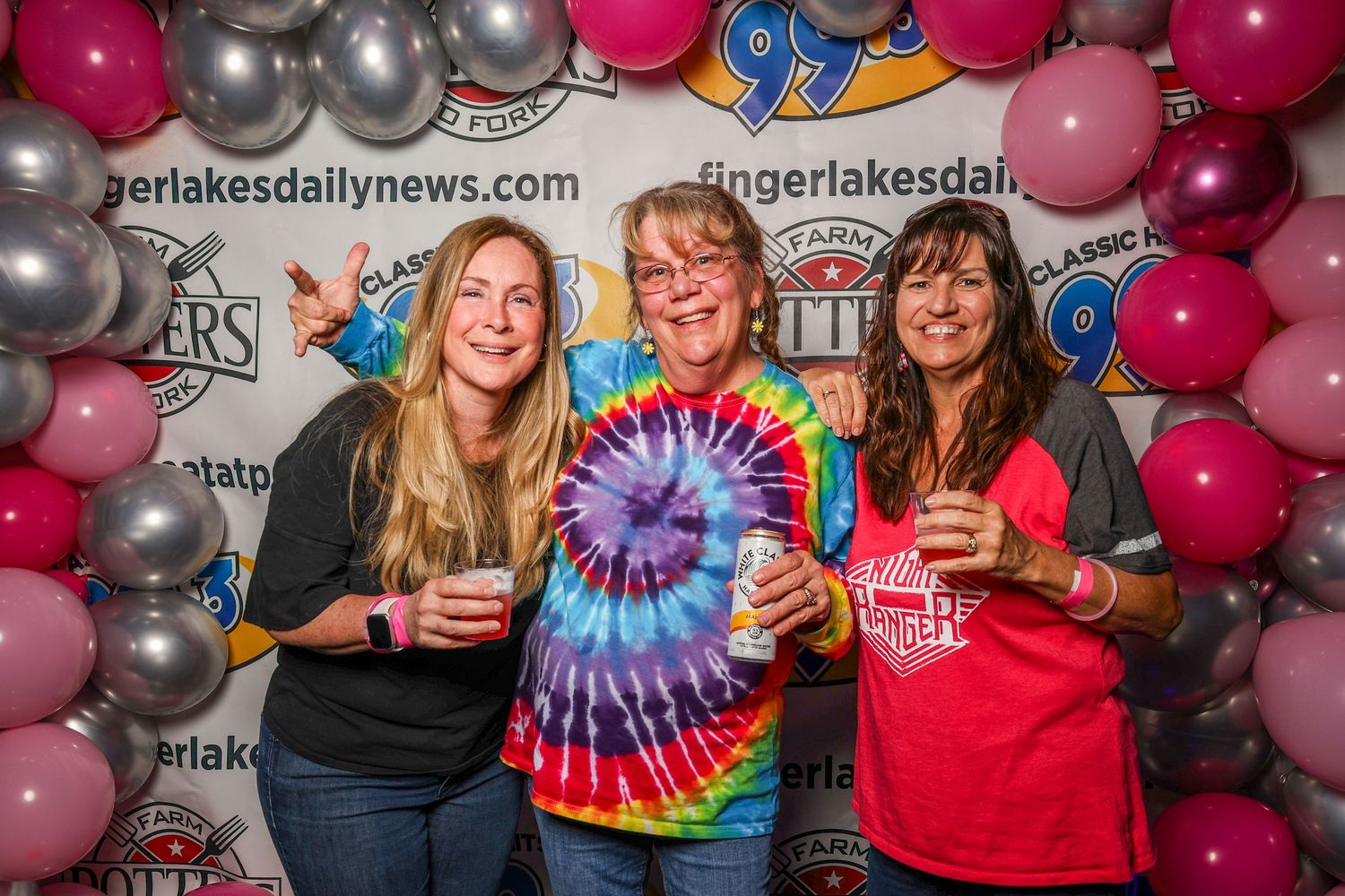 Three people pose together against a backdrop of pink, silver and red balloons while holding drinks and a tie-dye design.