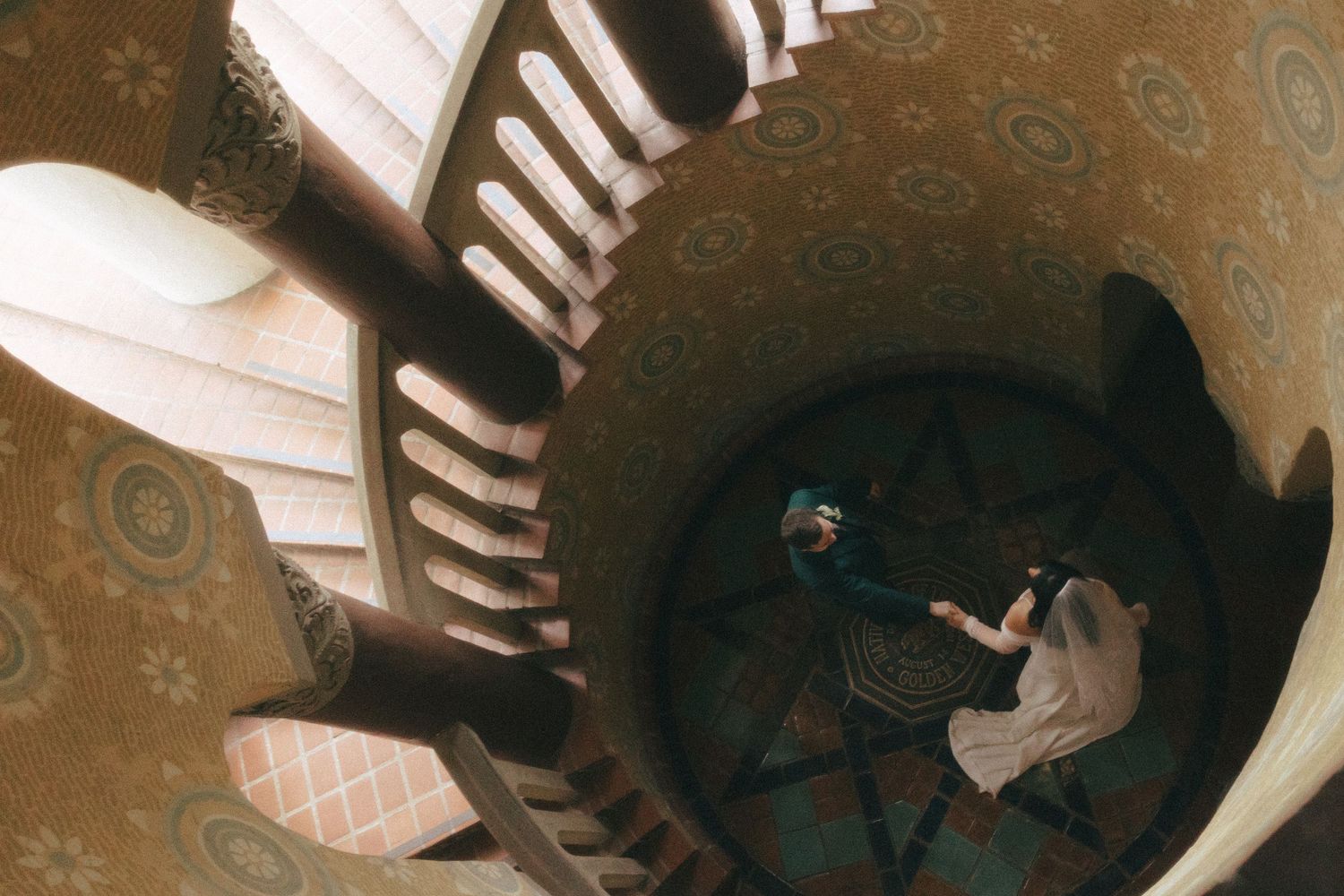 View looking down a spiral staircase with antique ornate wallpaper and a seated figure at the bottom.