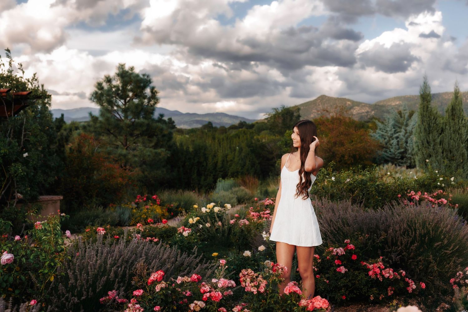 Young woman in white dress standing among colorful roses in a garden with mountain backdrop.