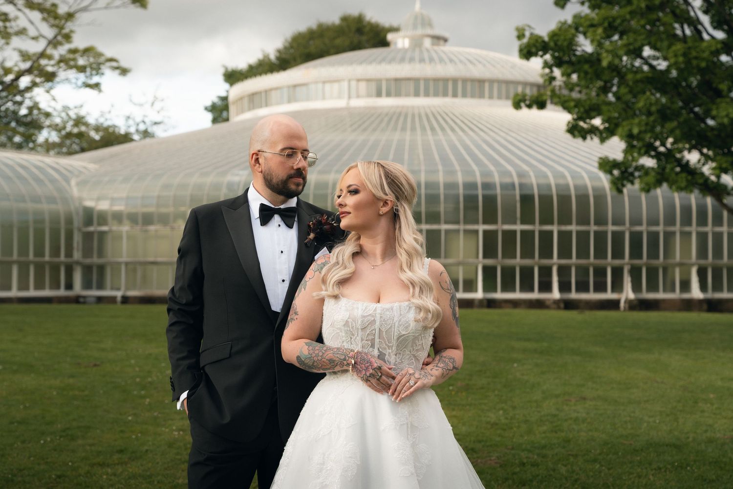 Bride and groom pose in front of a glass greenhouse, she in a strapless gown, he in a black tuxedo.