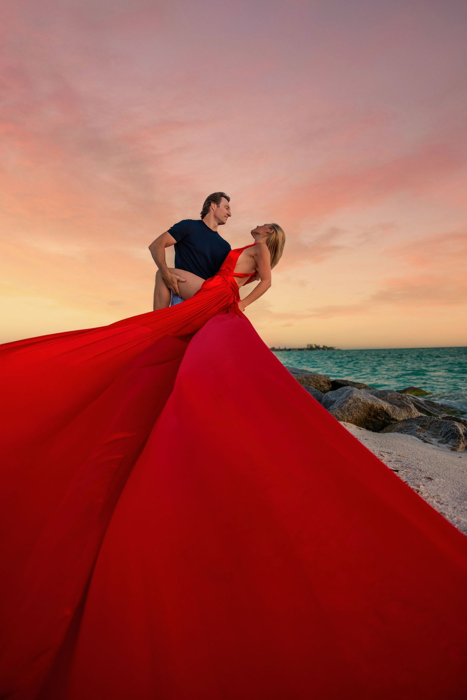 A couple poses against a stone wall as a flowing red gown creates dramatic movement in a series of romantic photos. Flying Dress Photoshoot