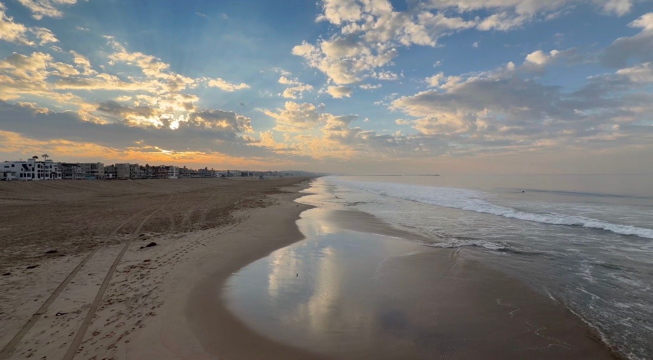 Panoramic sunset view of a sandy beach with dramatic clouds reflecting in the ocean waves and wet sand.