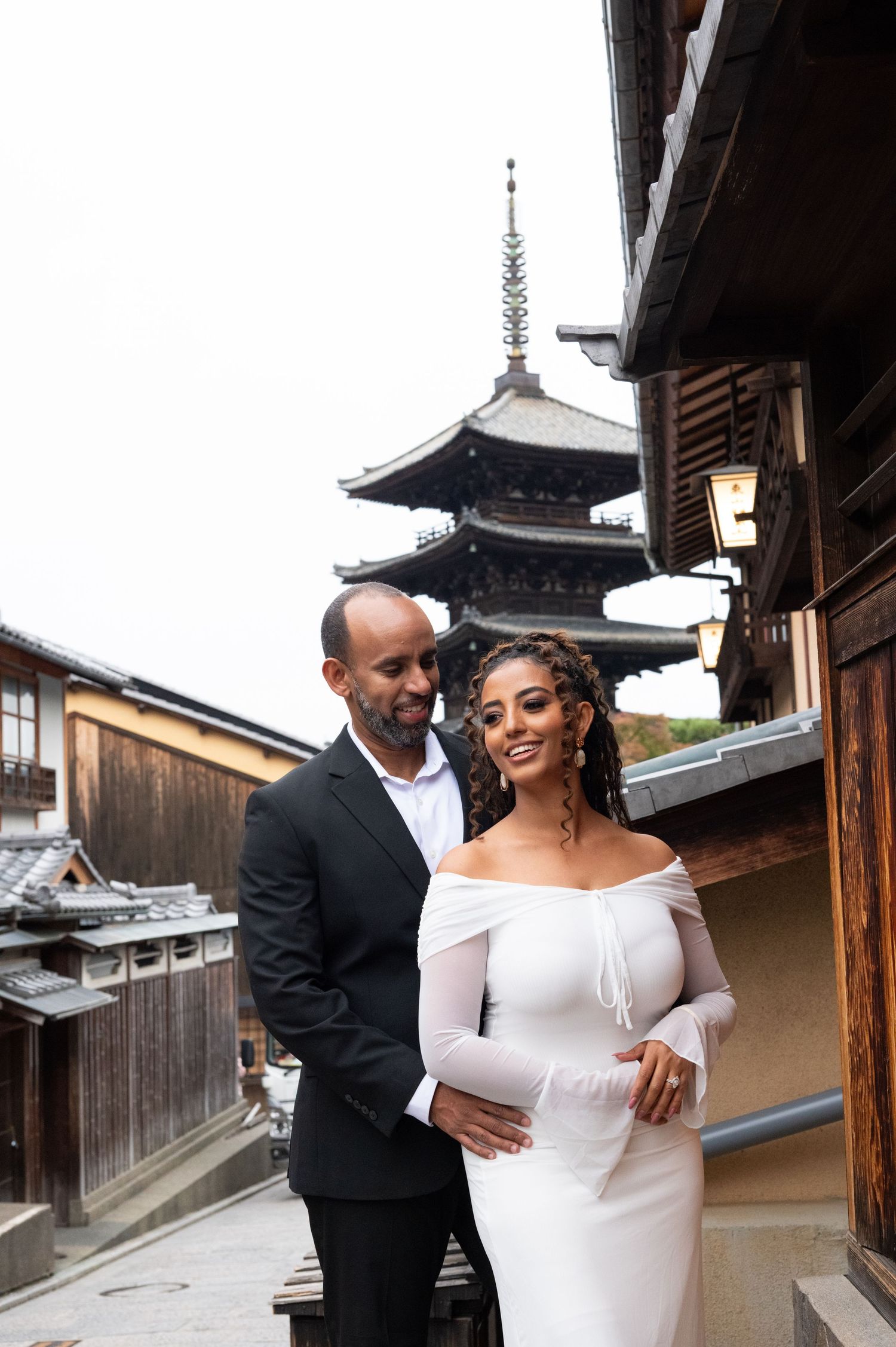 Elegant couple in formal wear posing near a traditional Japanese pagoda on a cloudy day.