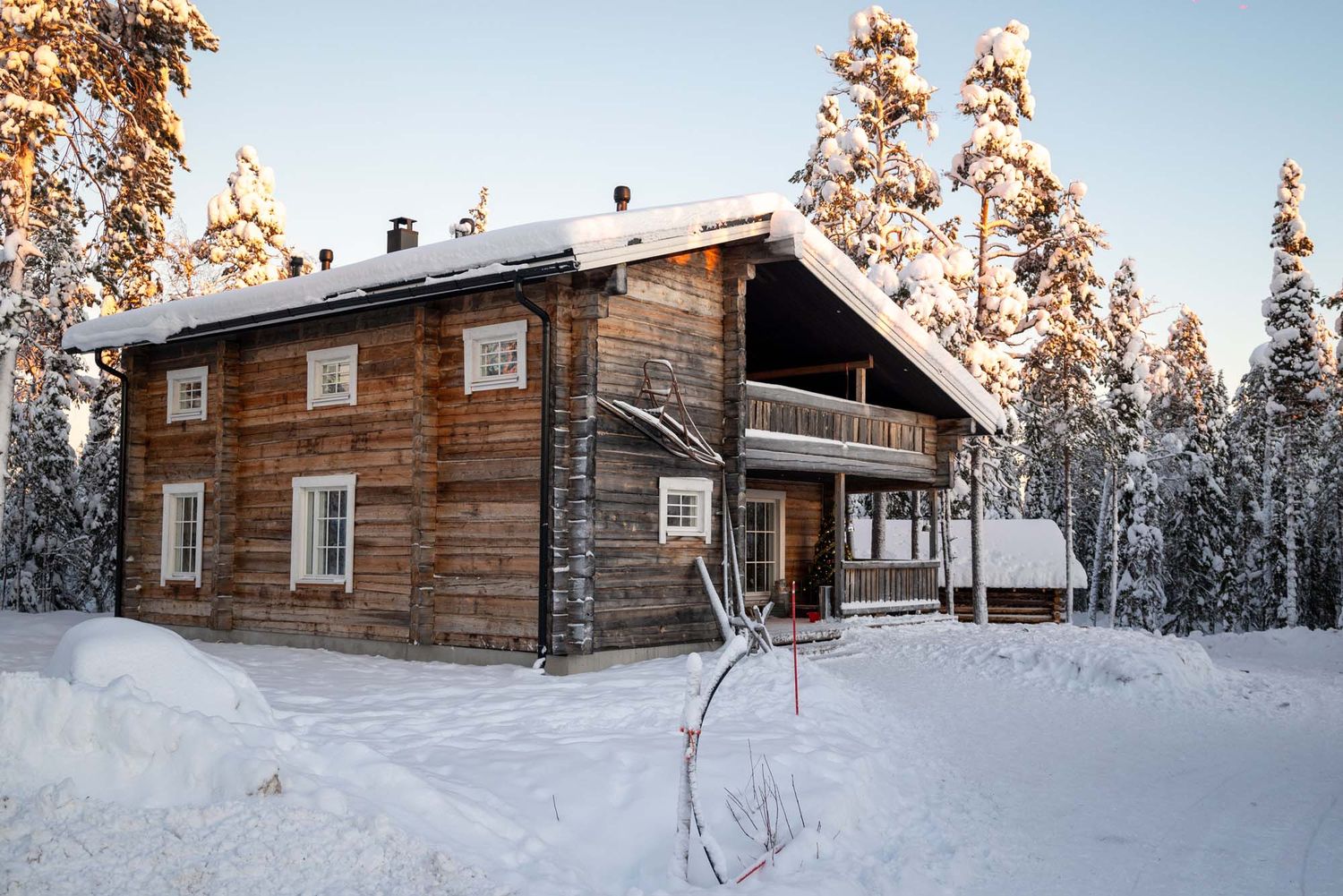Cozy log cabin surrounded by snowy pine trees in a winter forest landscape at sunset.