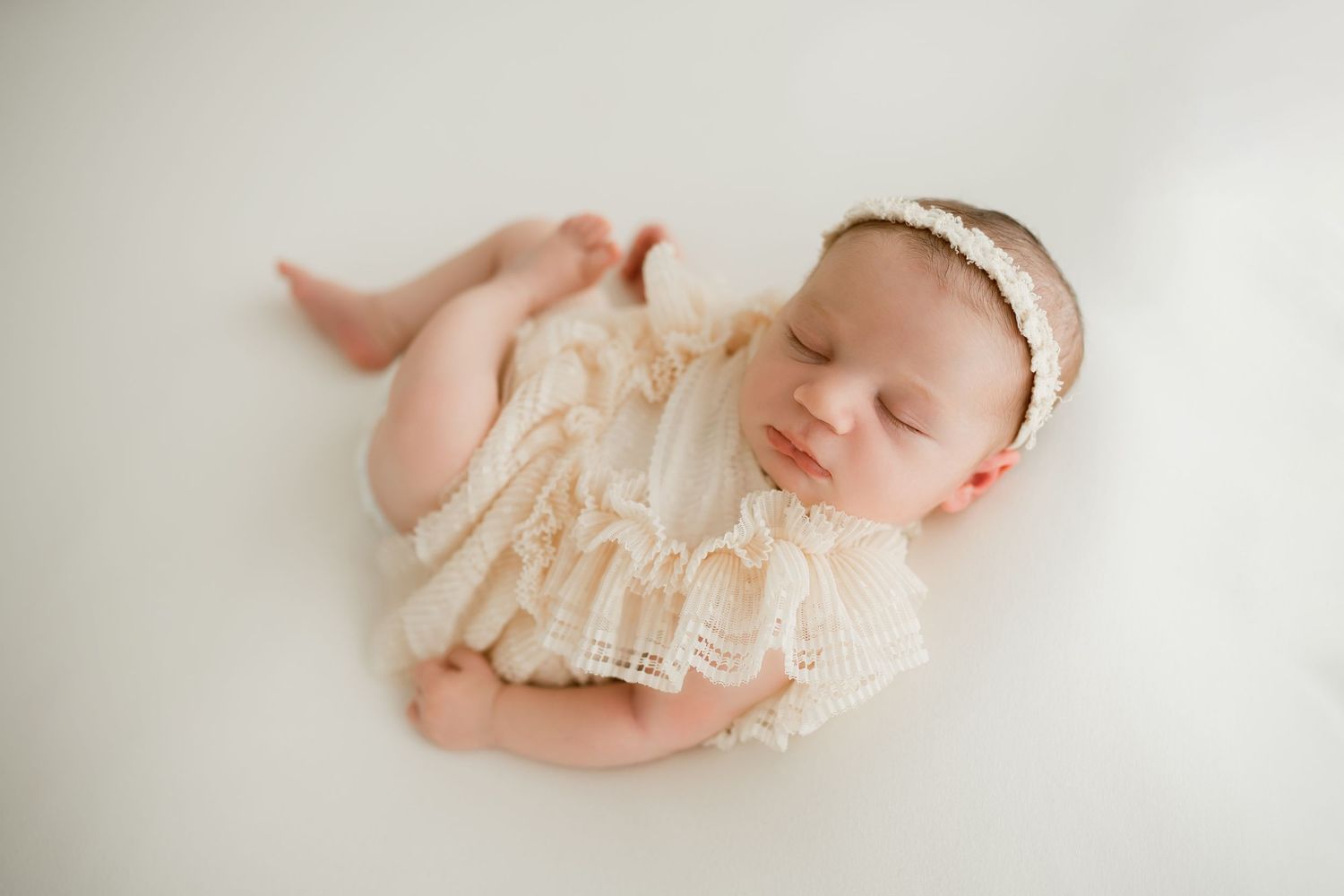 Sleeping newborn wearing delicate cream lace romper and pearl headband rests peacefully on white background.