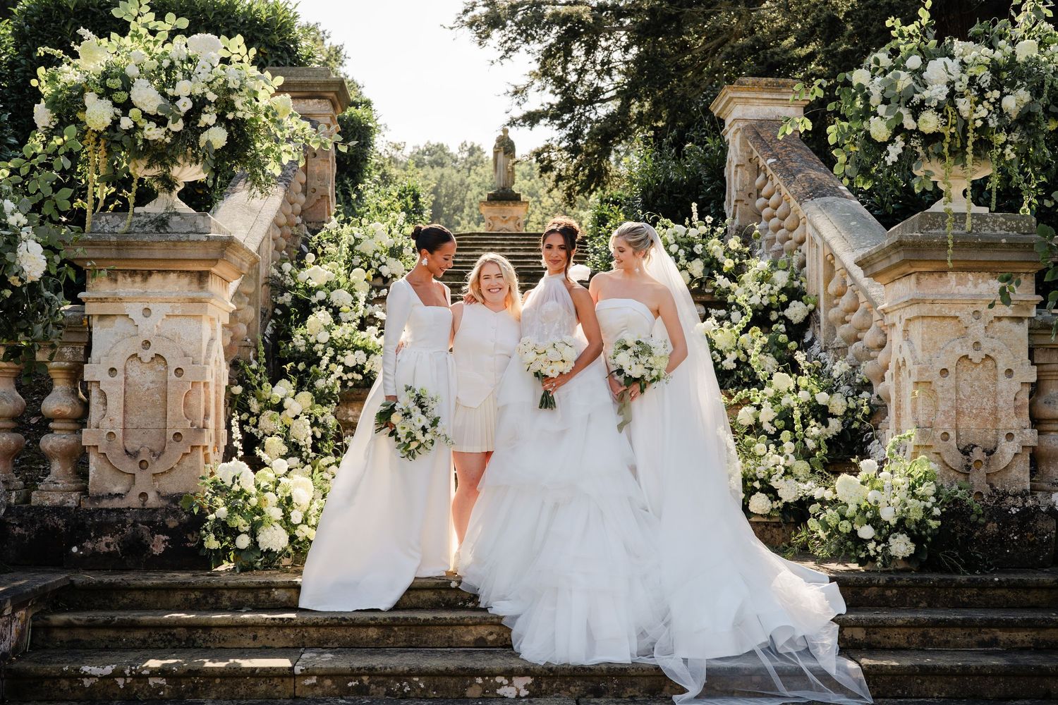 Wedding party in coordinated white outfits posed on garden stone steps.