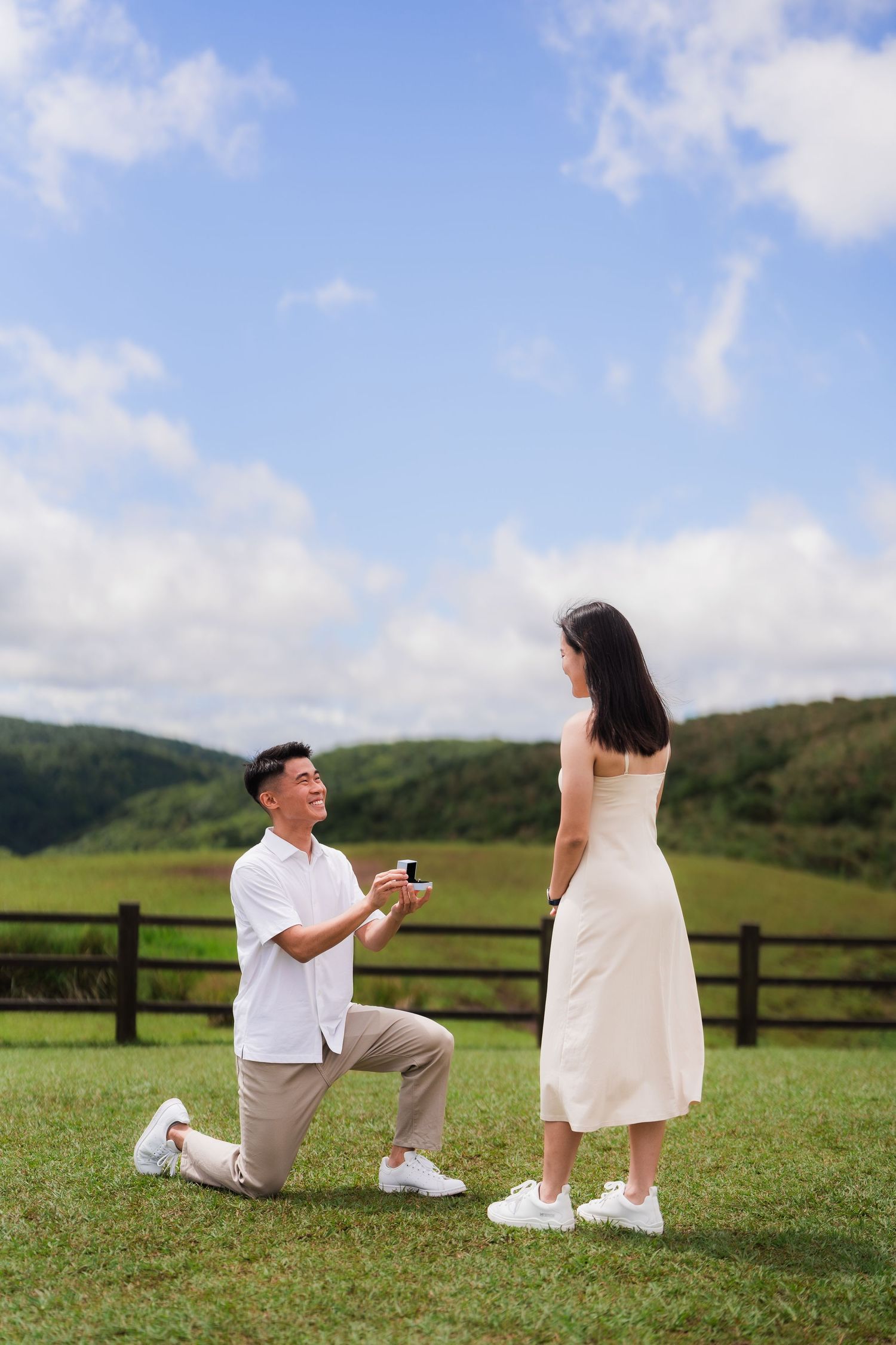 A romantic proposal moment on a grassy hilltop with wooden fences and blue skies in the background.