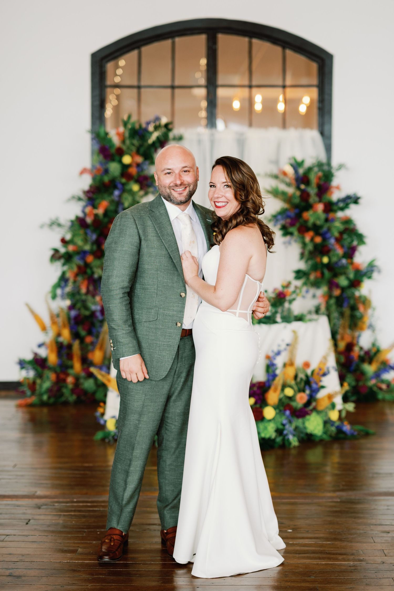 A couple poses in front of a floral arch with greenery and colorful flowers against an arched window backdrop.