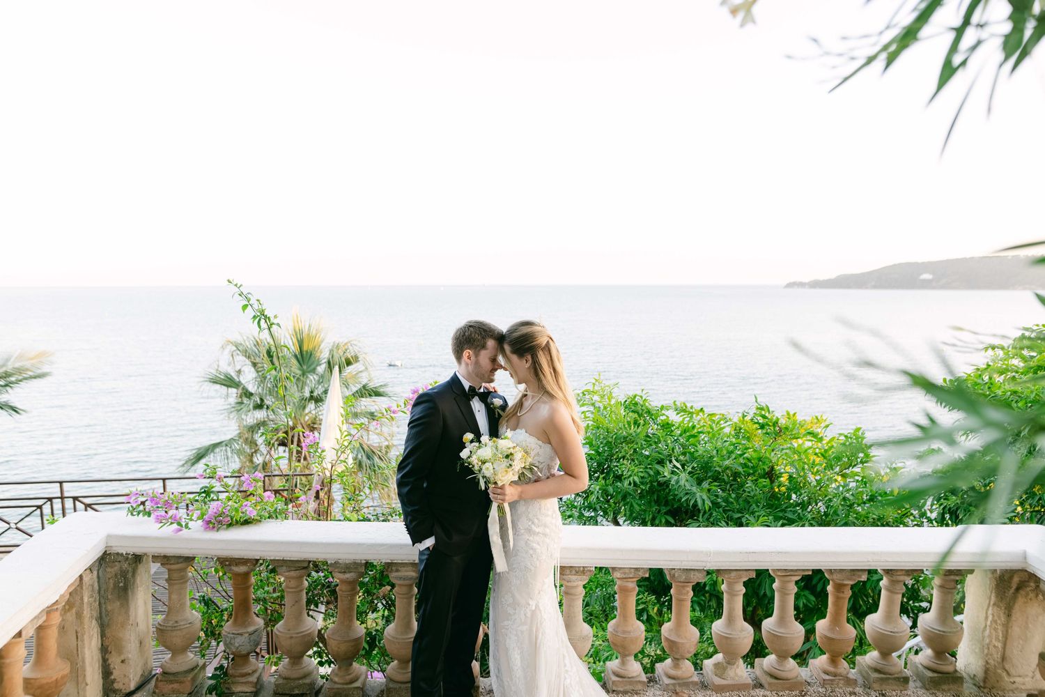 A couple shares a romantic moment during their outdoor wedding ceremony at a scenic stone balcony overlooking gardens.