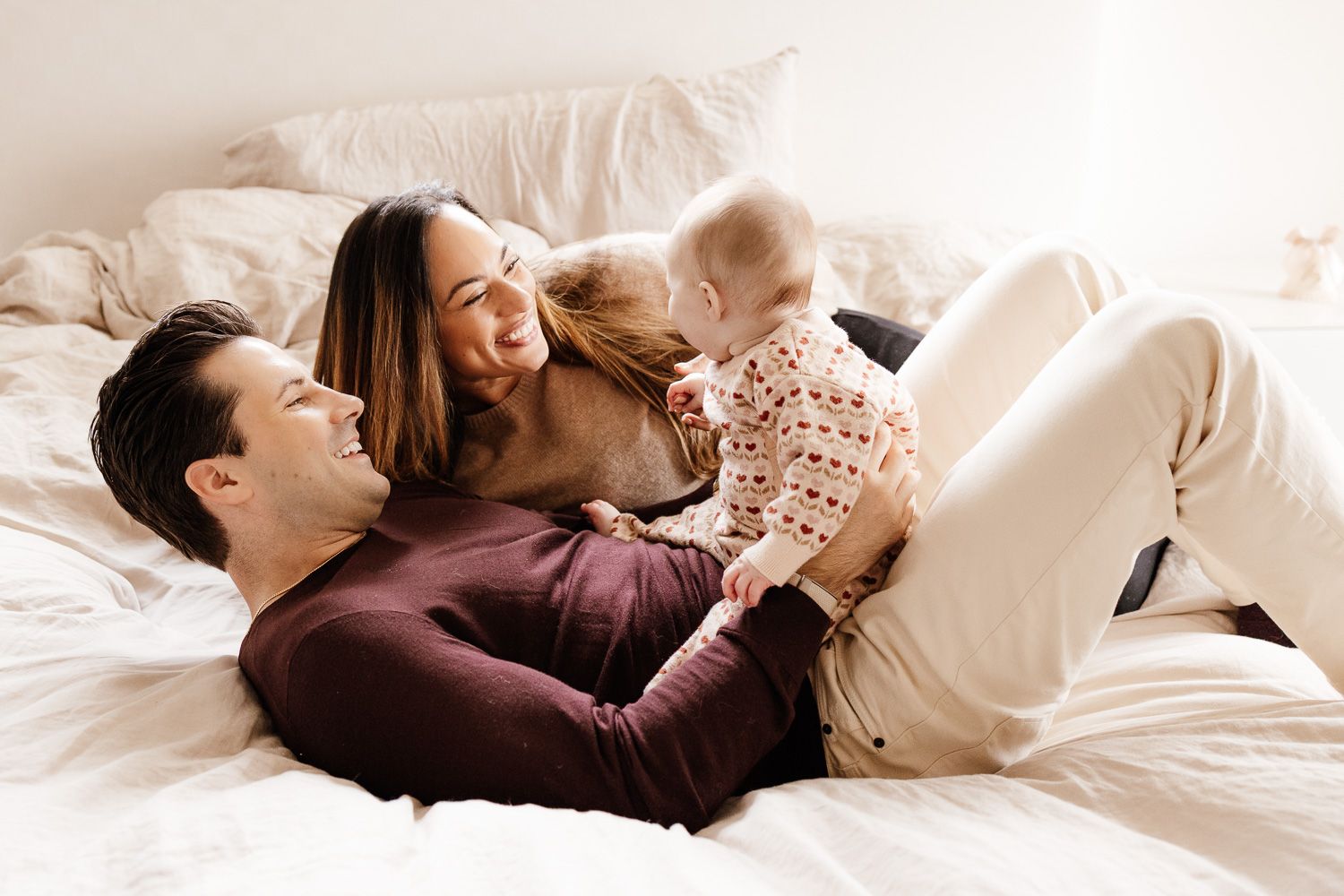 Couple lying together on their white bed while holding their six-month old baby during their milestone photography session