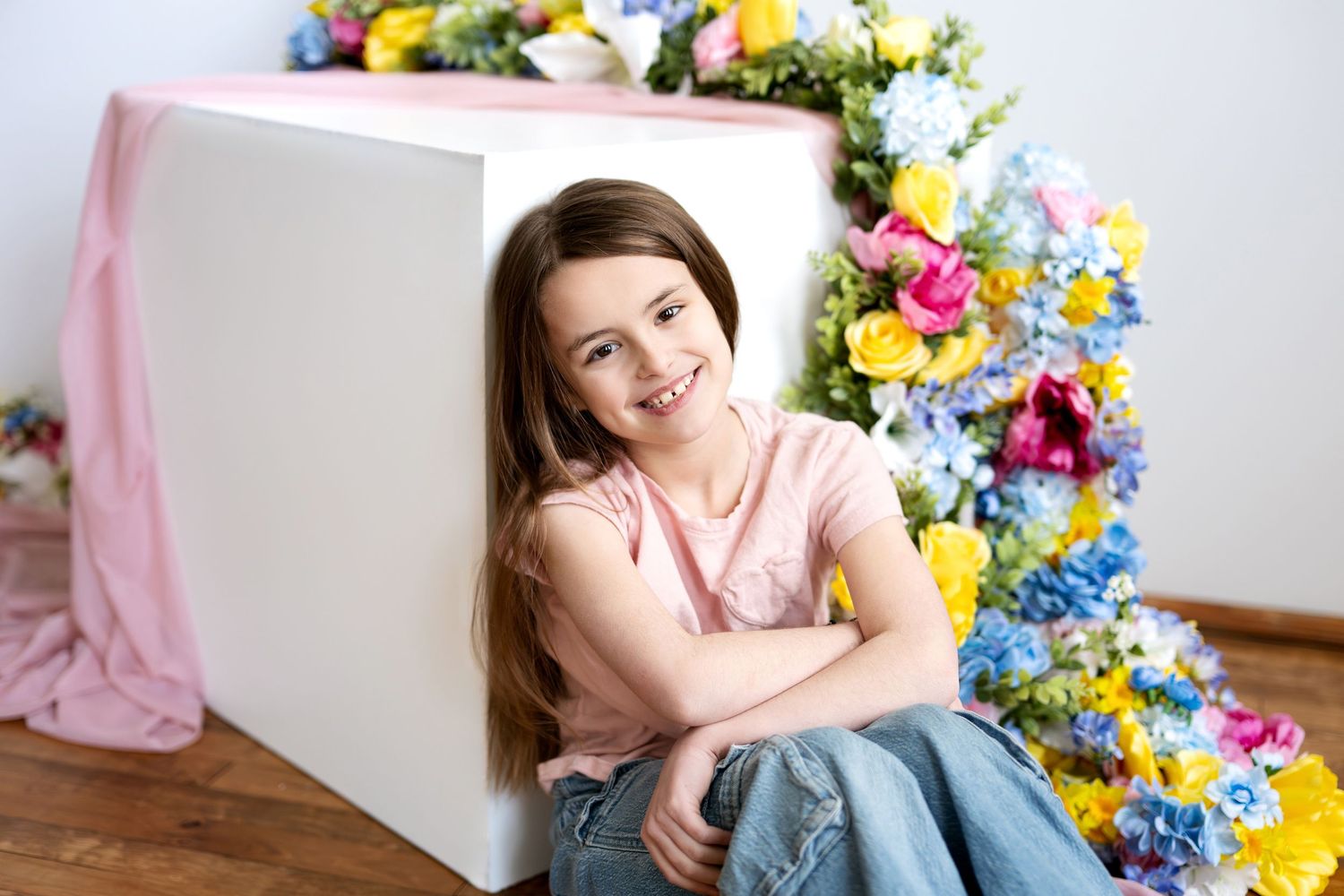 Smiling young girl with long brown hair sitting against white cube with colorful spring flowers.