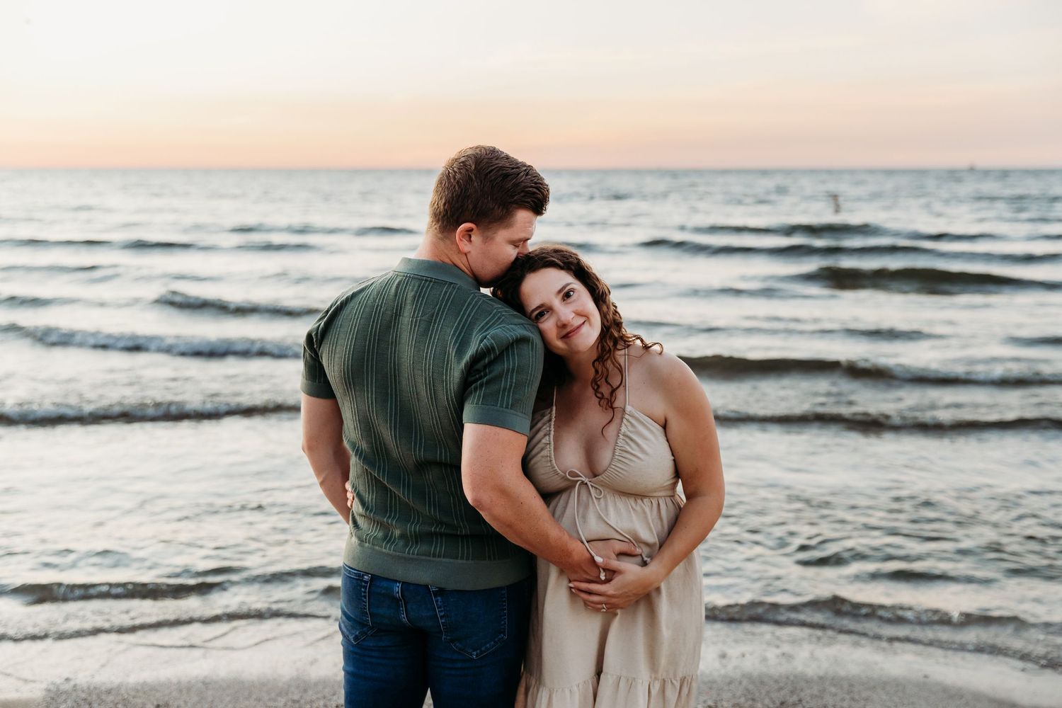 A couple embraces on a sandy beach at sunset with gentle waves rolling in along the shoreline. Cleveland Maternity Photographer