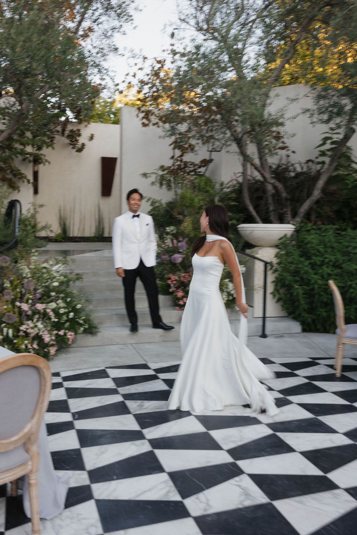 A couple dances together on a striking black and white checkered outdoor patio surrounded by lush greenery and white walls.