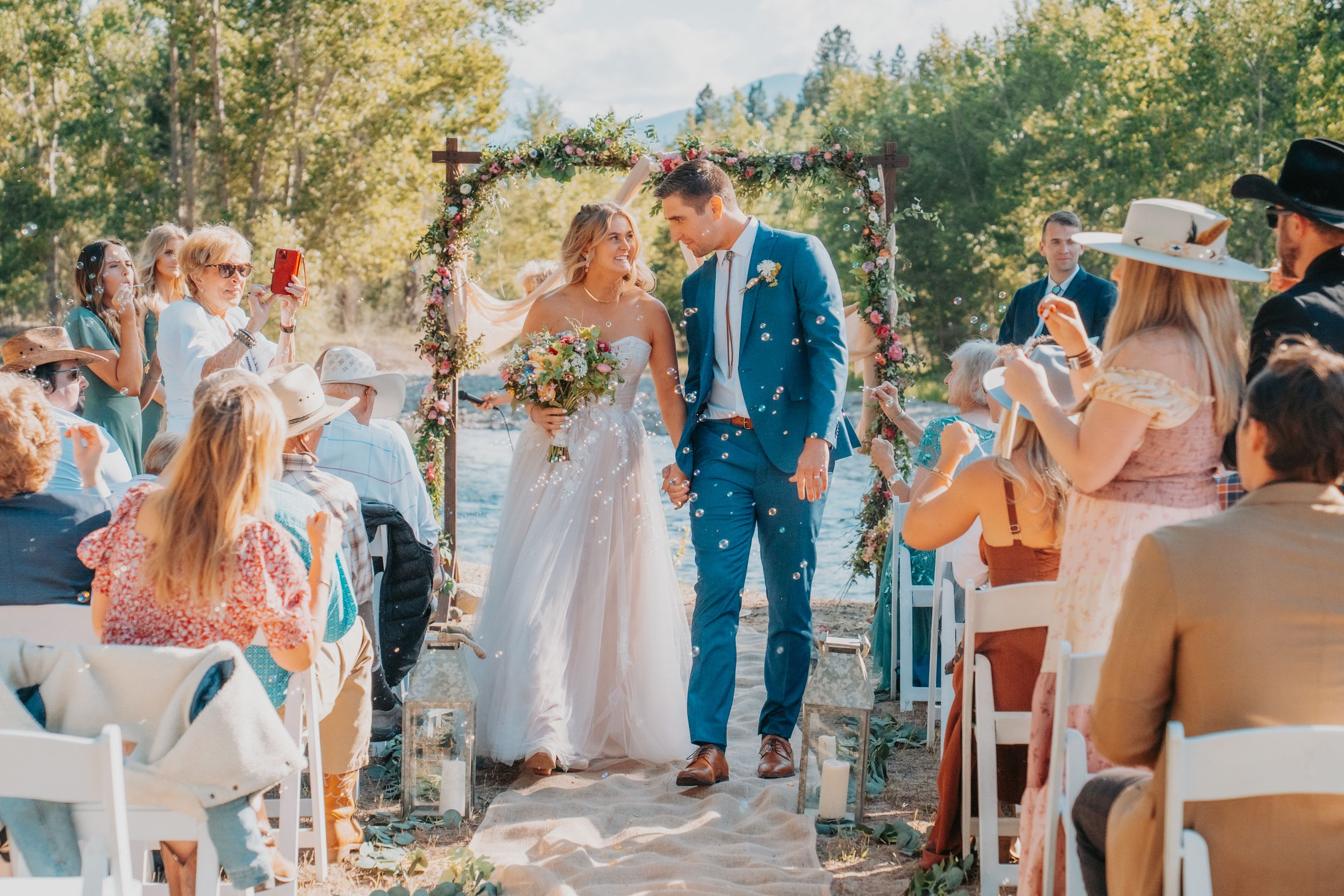 A sunny outdoor wedding ceremony with guests seated on white chairs as a couple walks down the rustic wooden aisle.