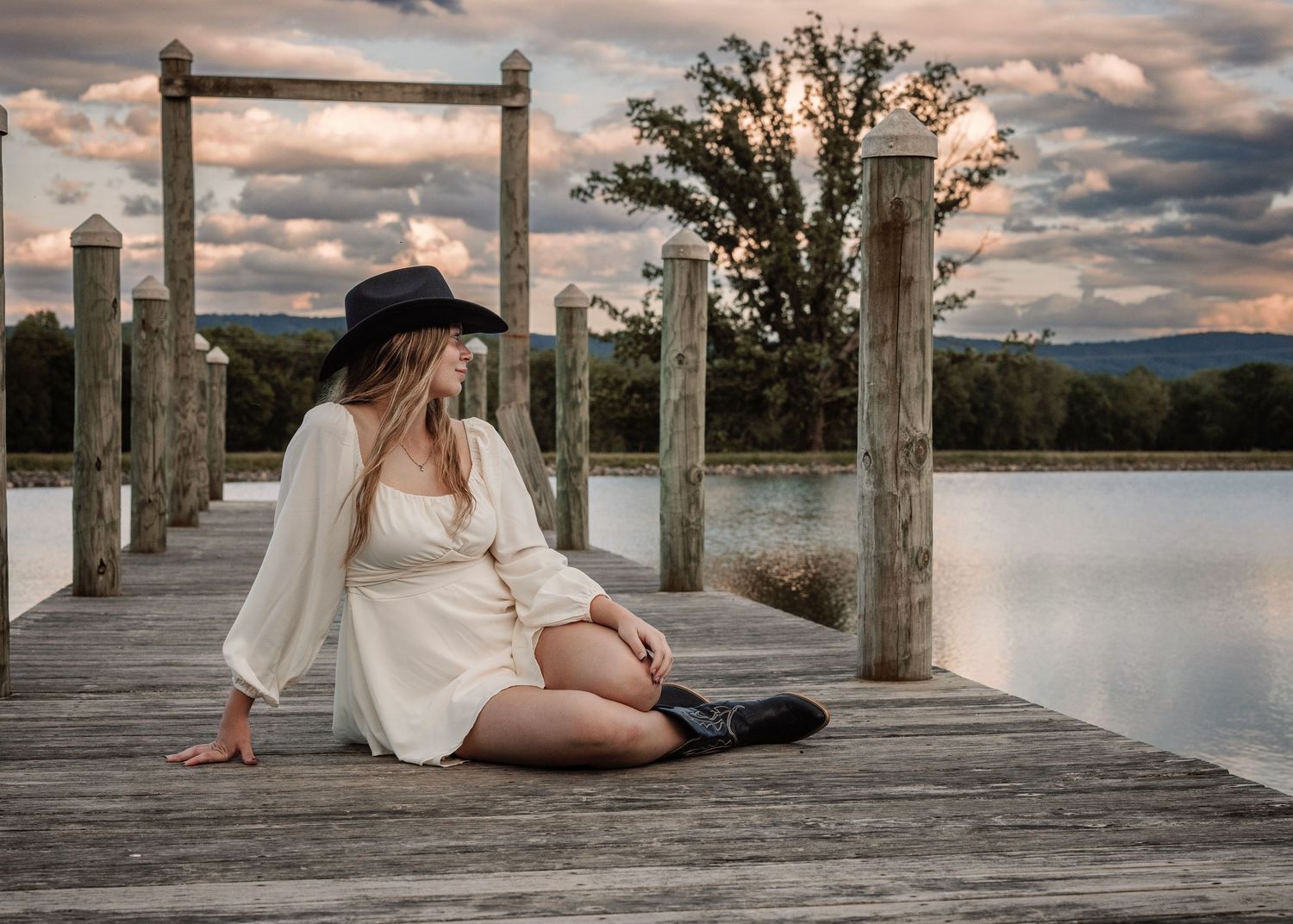 Person in white dress relaxing on wooden dock at sunset over calm waters.
