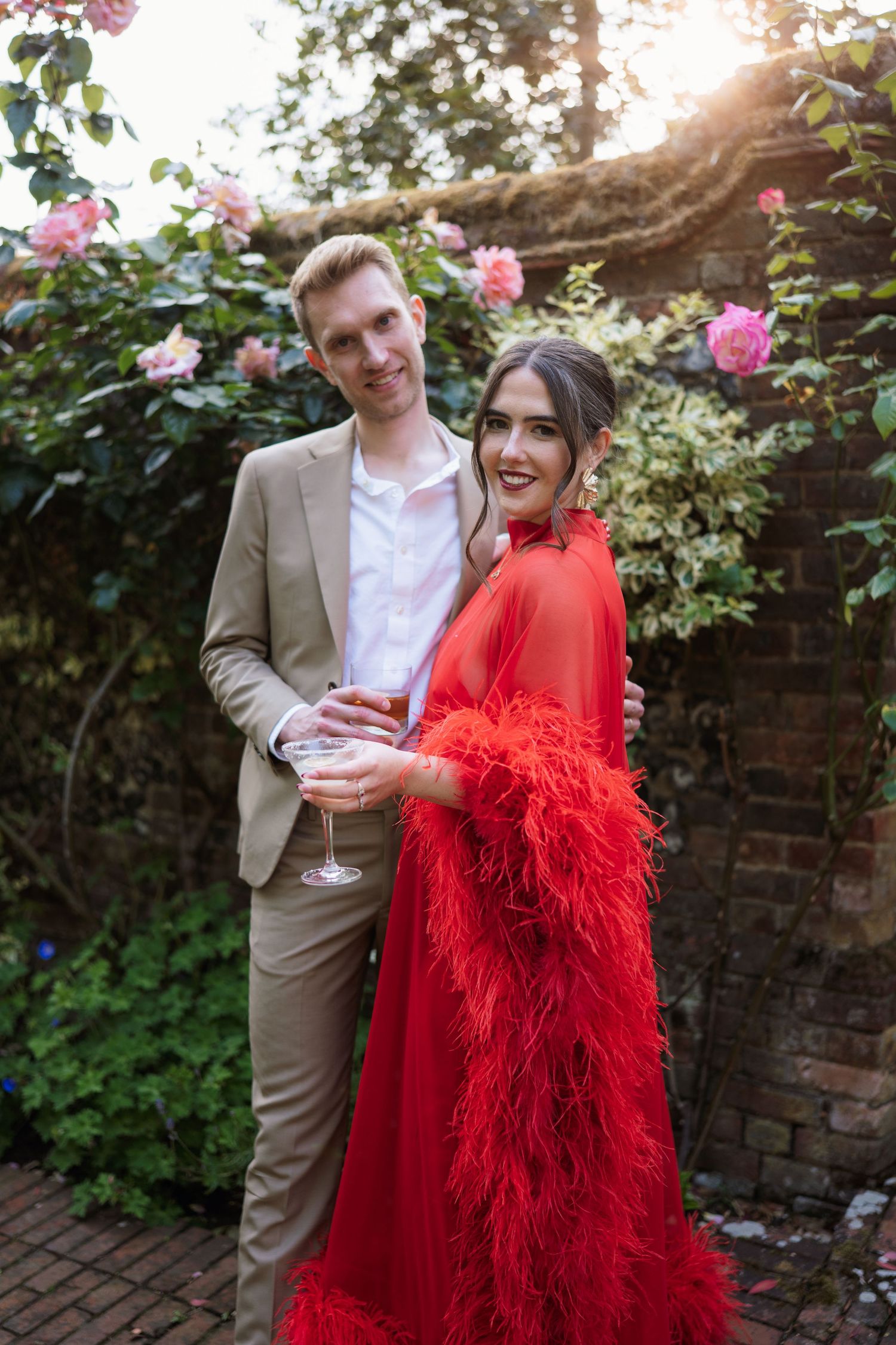 A couple in formal attire shares a romantic moment in a garden with pink roses and sunlight peeking through.