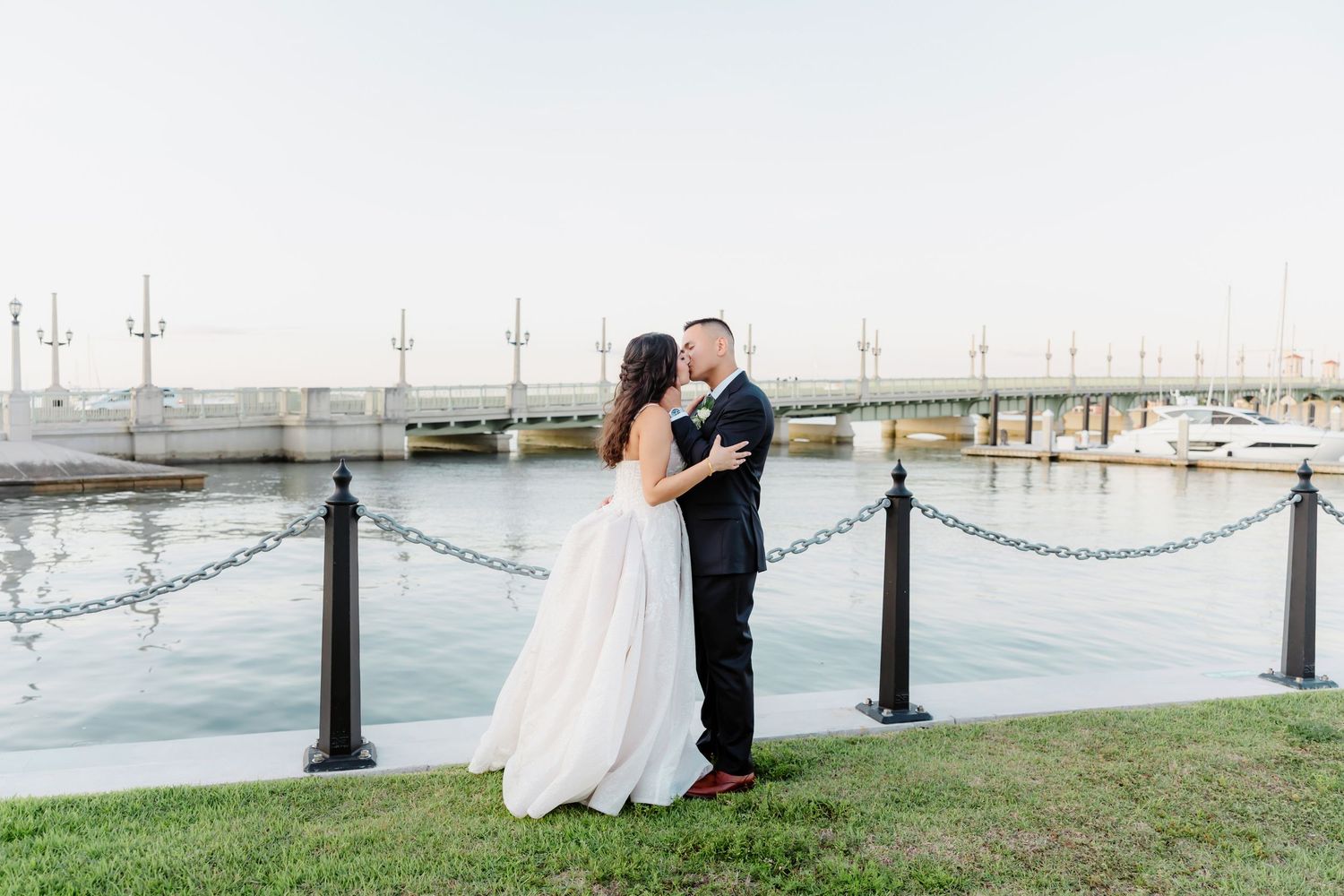 A couple shares a romantic moment by the waterfront with boats docked at a marina in the background.
