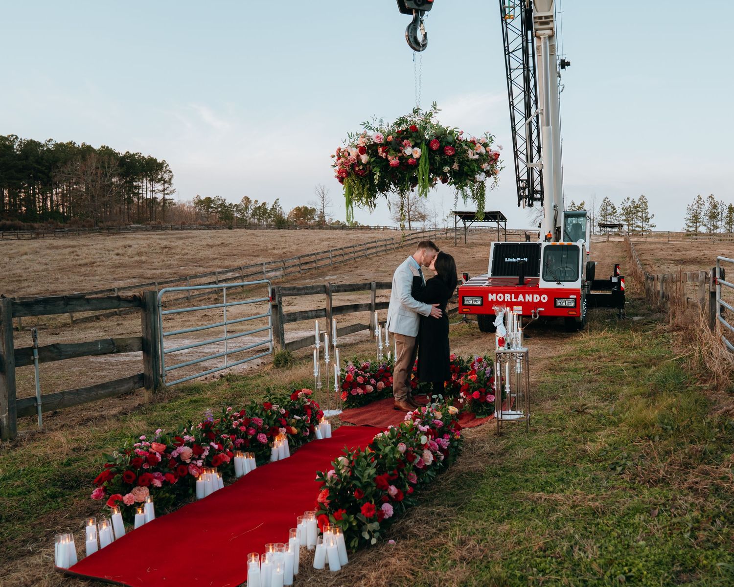 A romantic outdoor proposal setup with red carpet, white candles, and floral arrangements beneath a tall crane.
