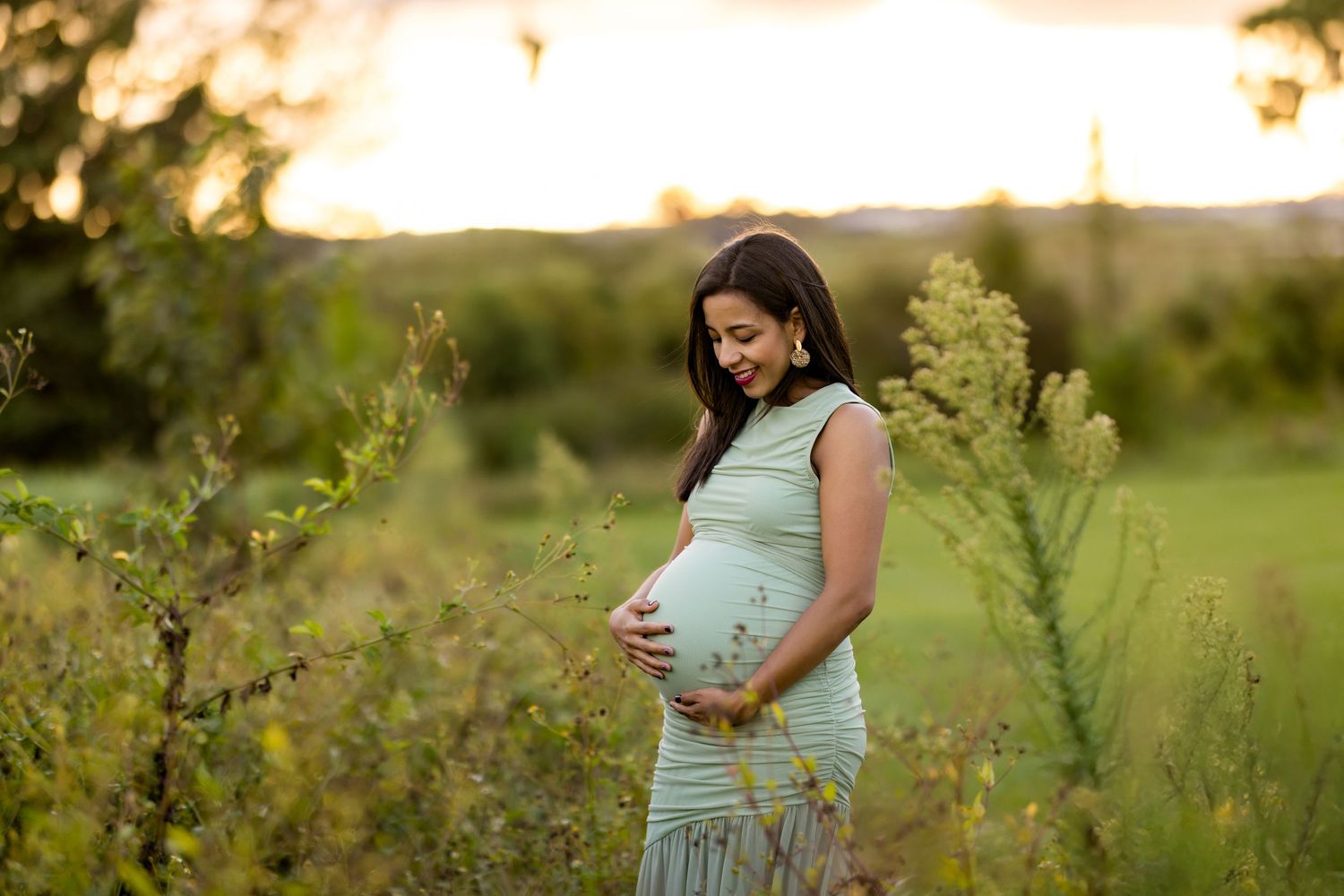 A pregnant woman in a mint green dress stands in a sunlit meadow during golden hour.