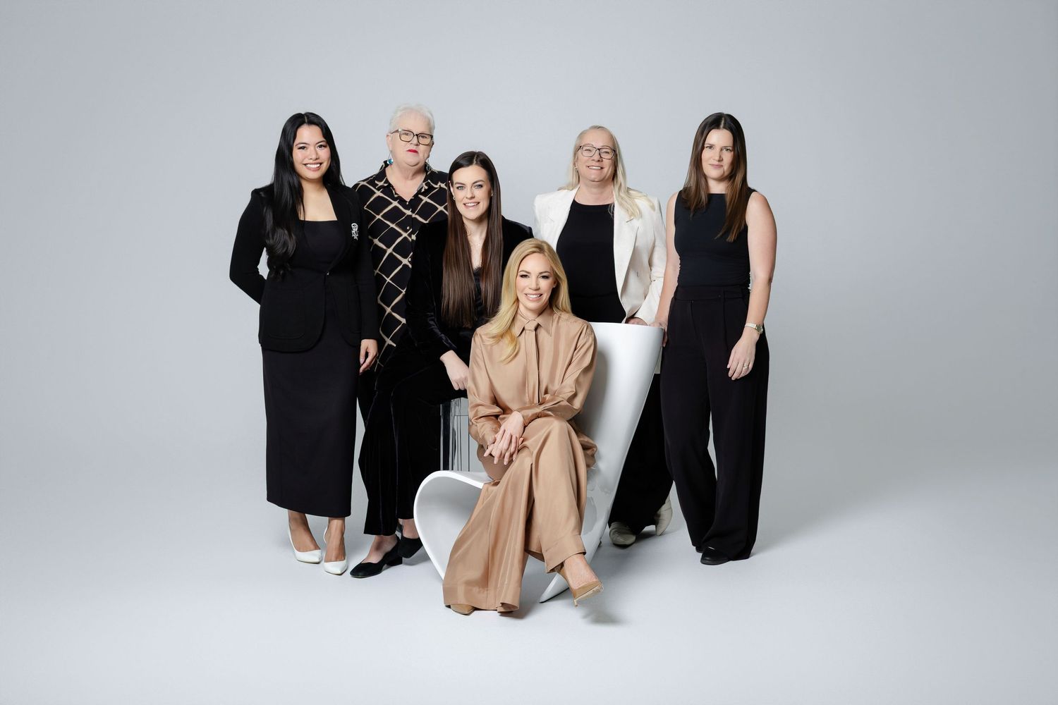 Group of six people wearing elegant black outfits and one in beige posing together against white background in studio setting.