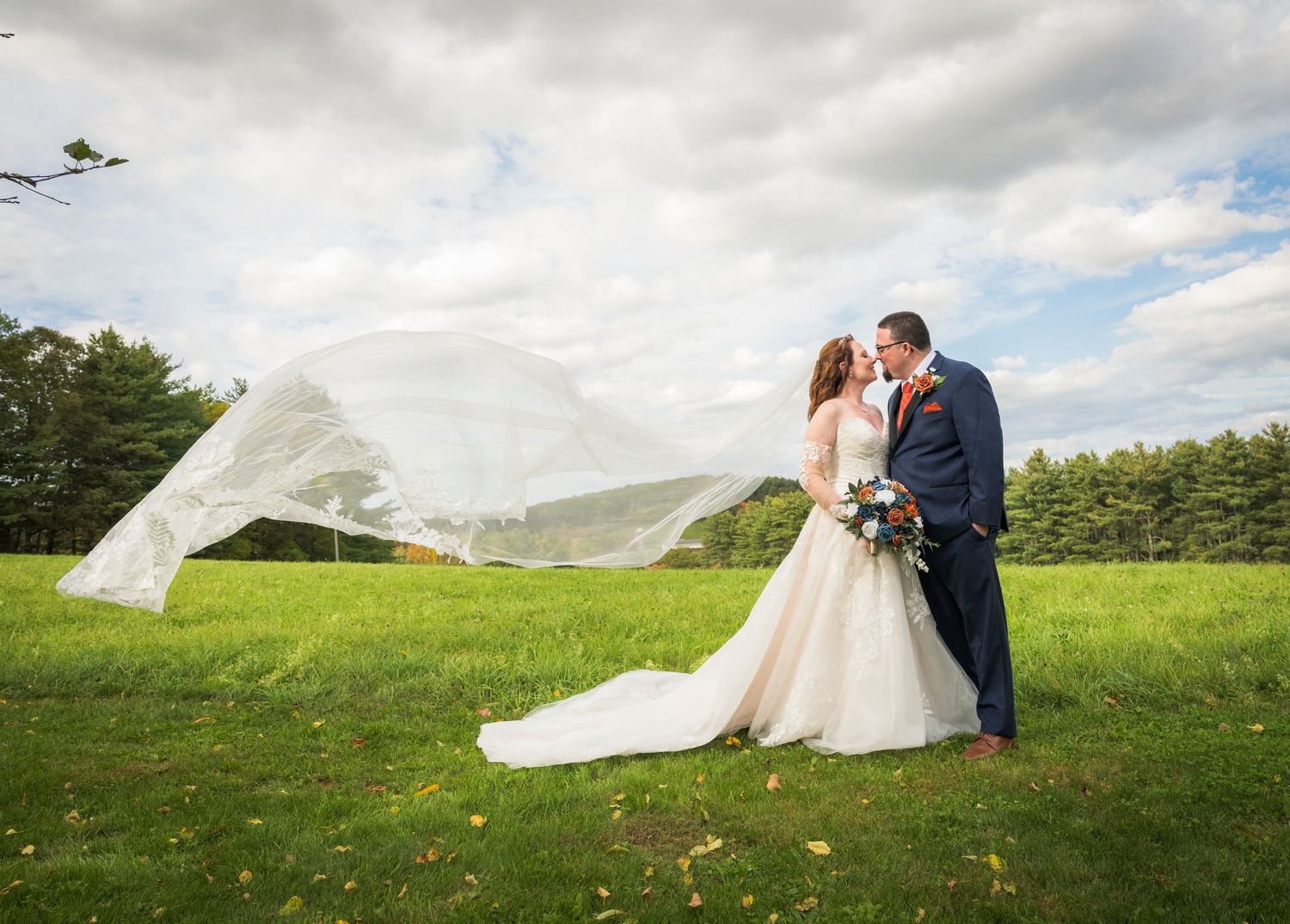 Newlyweds share an intimate moment outdoors with autumn foliage and rustic architecture in background.