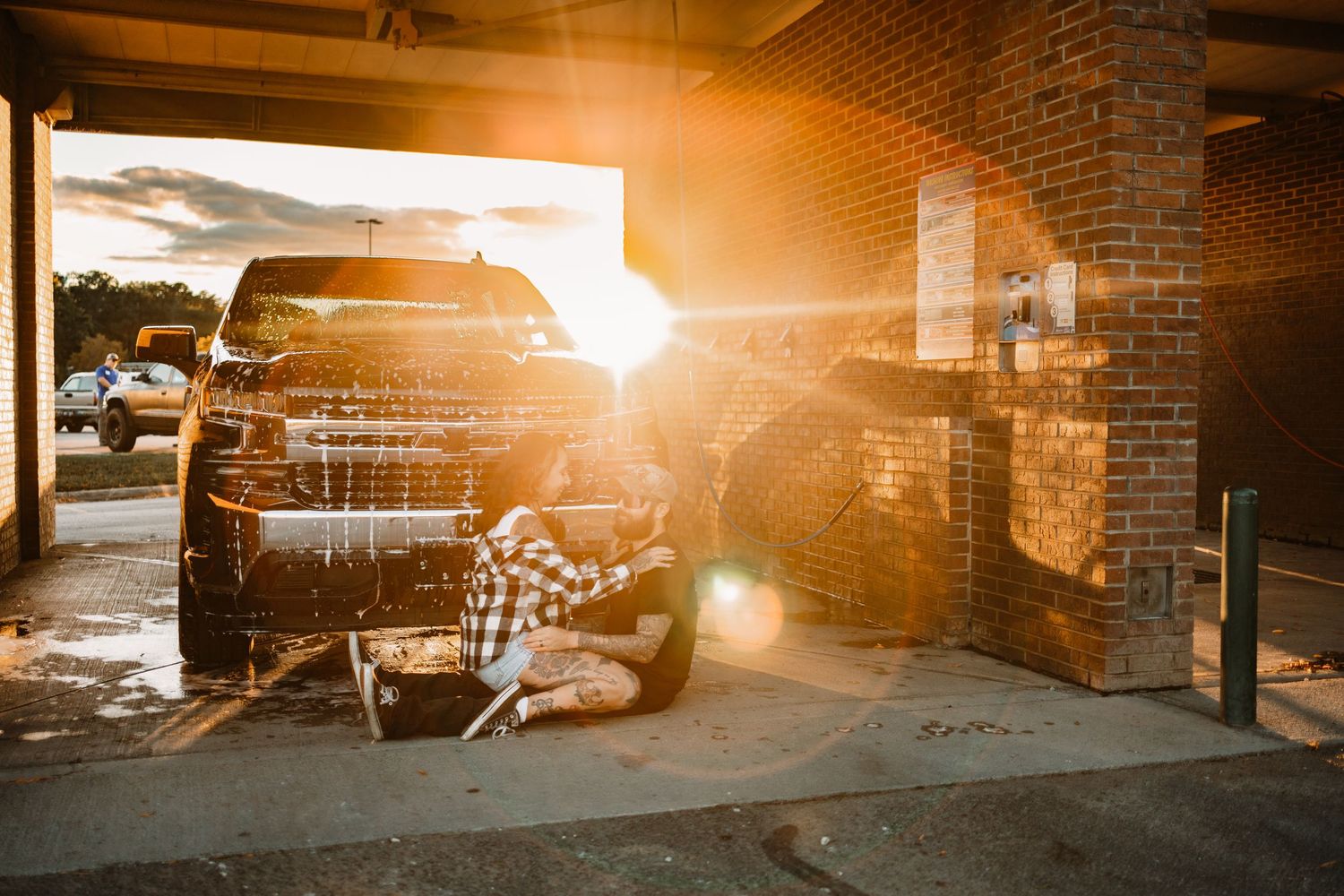 Person washing black truck at outdoor car wash bay with dramatic golden sunset lens flare.