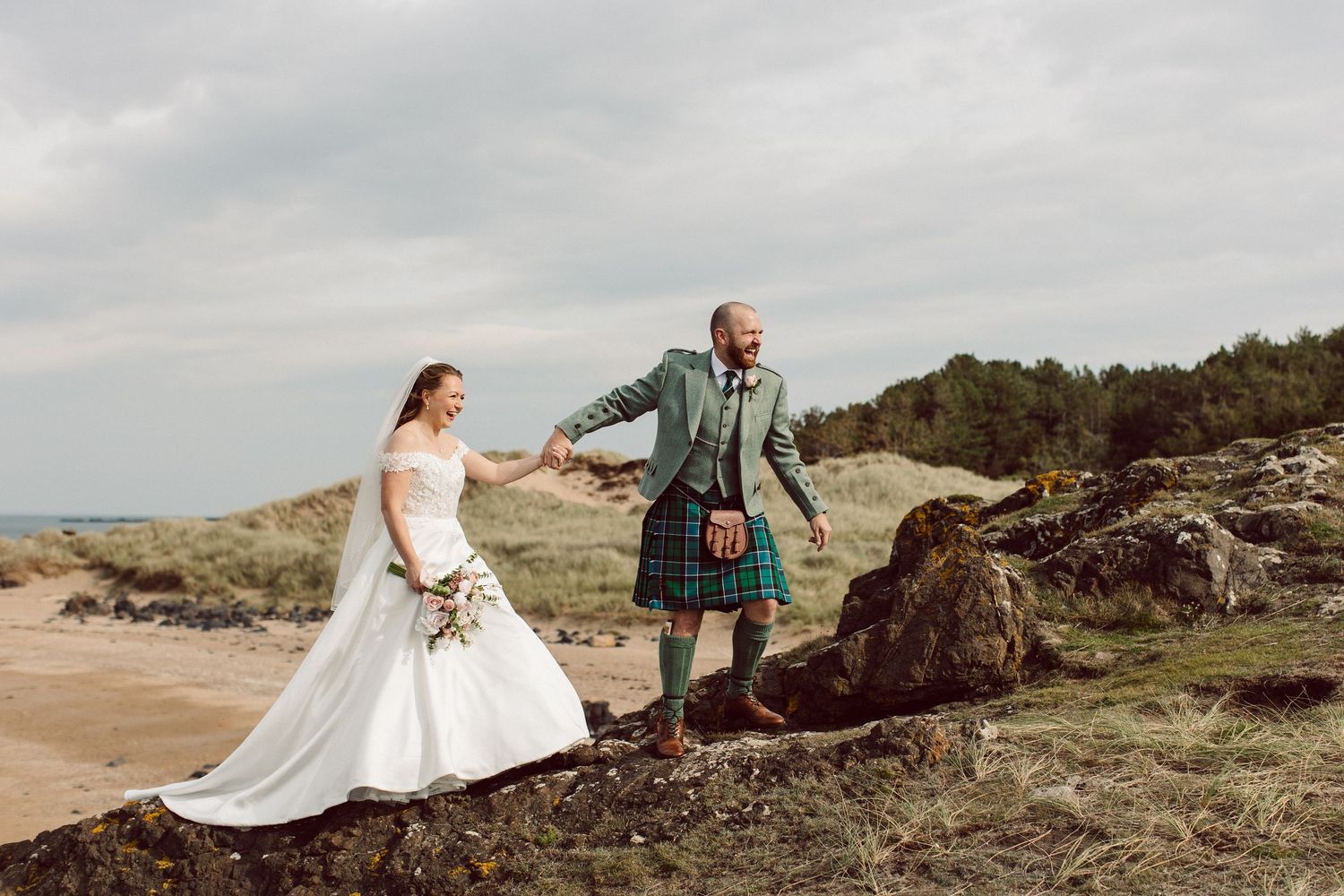 A couple in wedding attire stands on a rocky coastal cliff, with the groom wearing a traditional Scottish kilt.