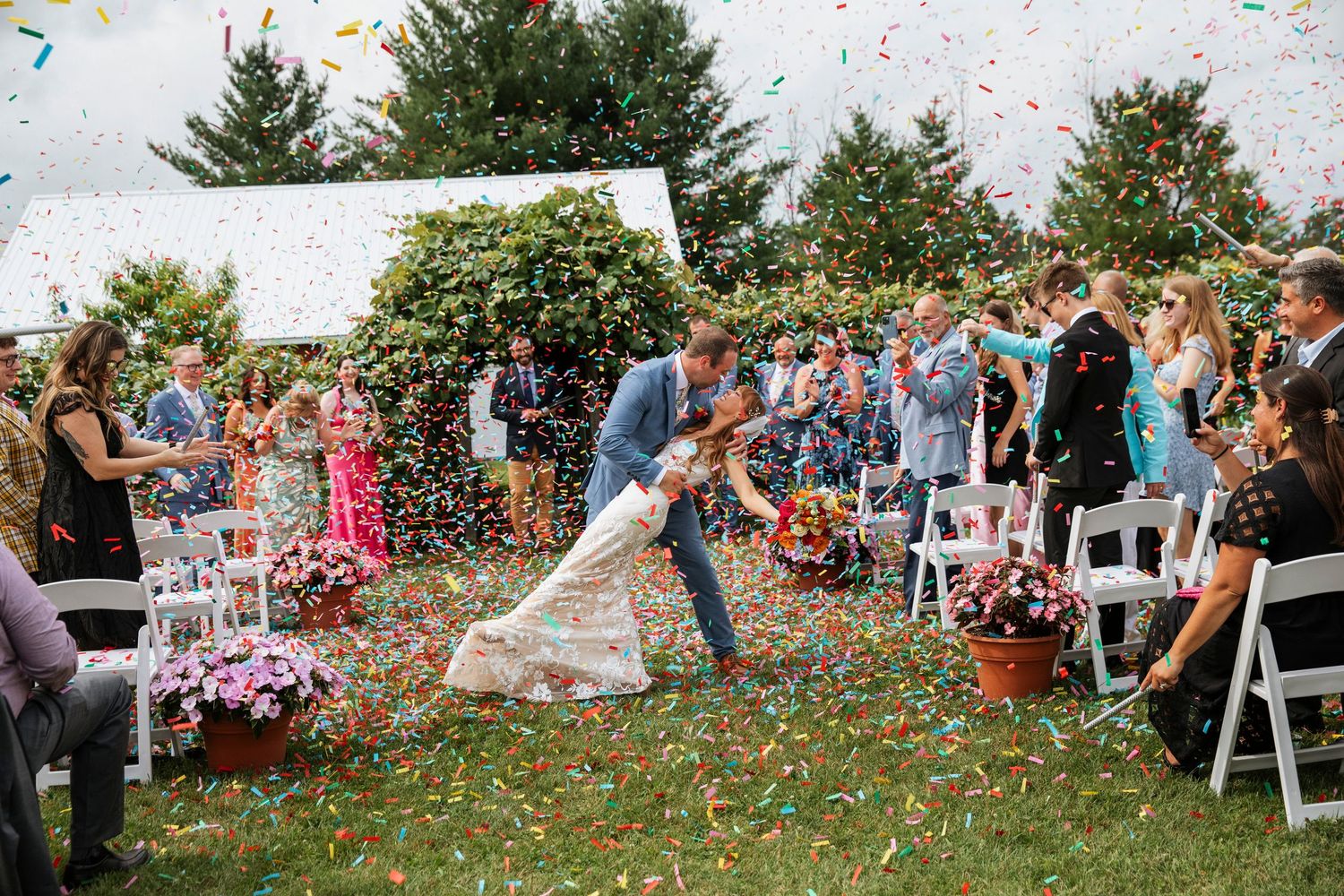 Bride and groom share a romantic dip kiss while colorful confetti falls during outdoor wedding ceremony.
