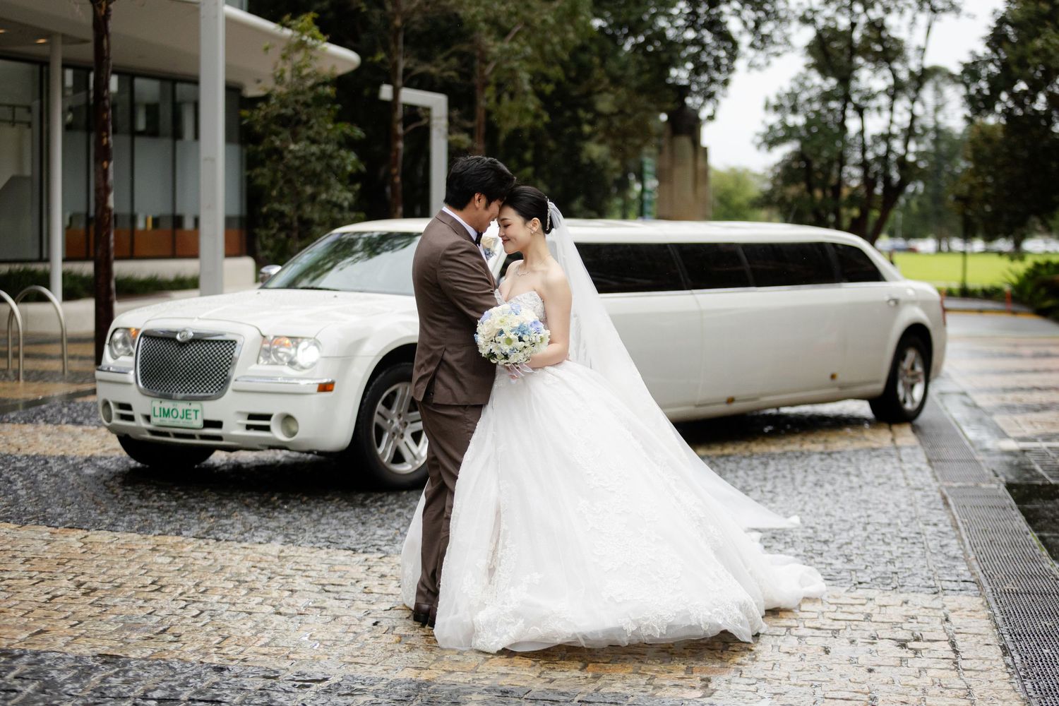 A newlywed couple walks along a charming cobblestone path on a rainy day near parked vintage automobiles.
