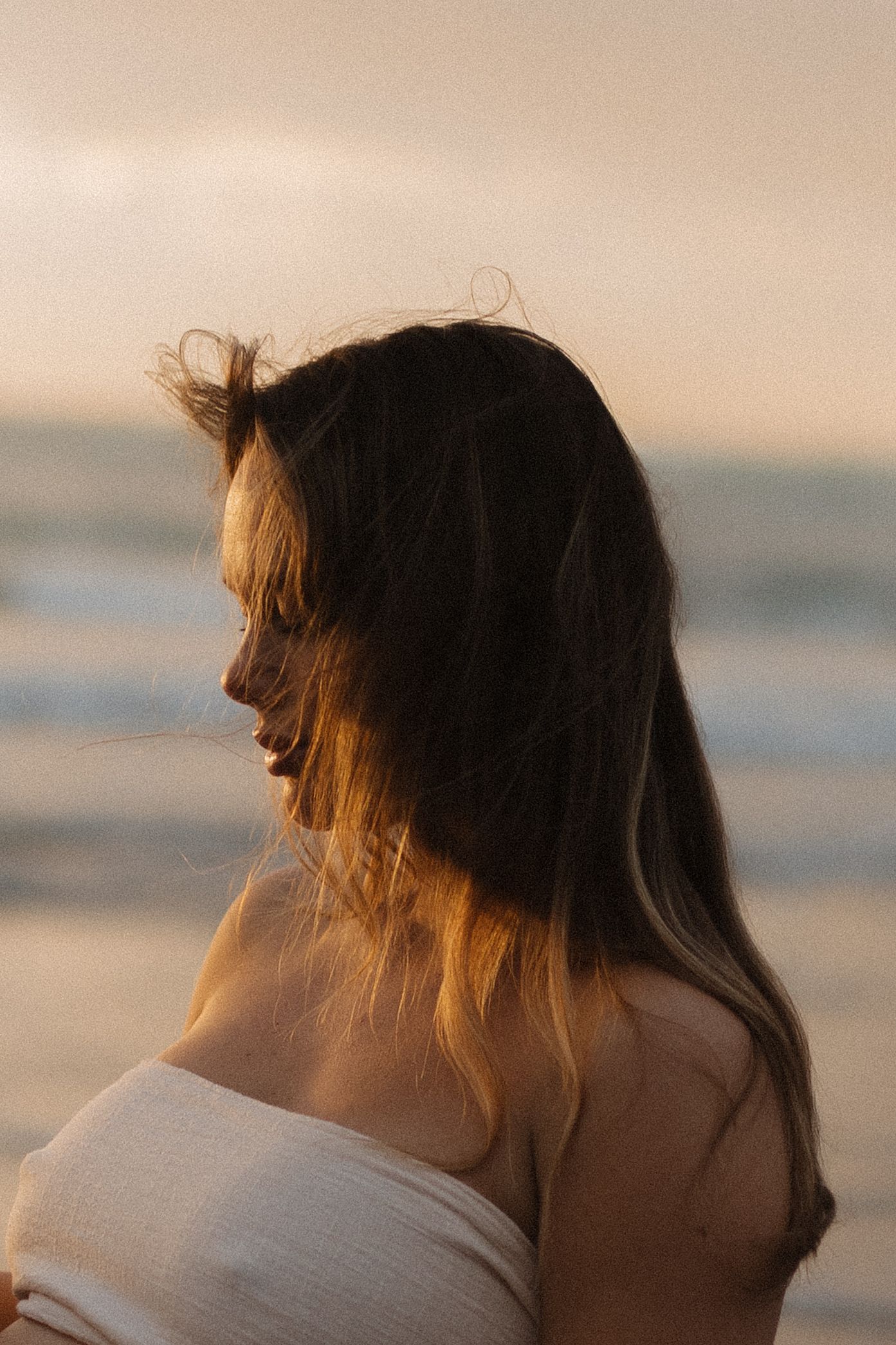 A pregnant woman in white dress stands on beach at sunset, silhouetted against golden sky.