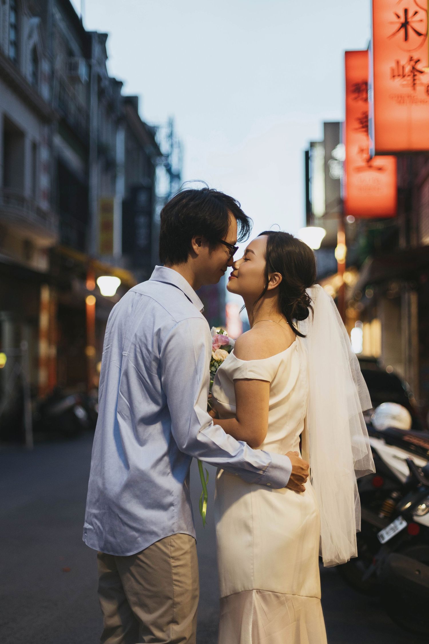 Asian newlywed couple sharing romantic moment on city street at twilight with neon signs.